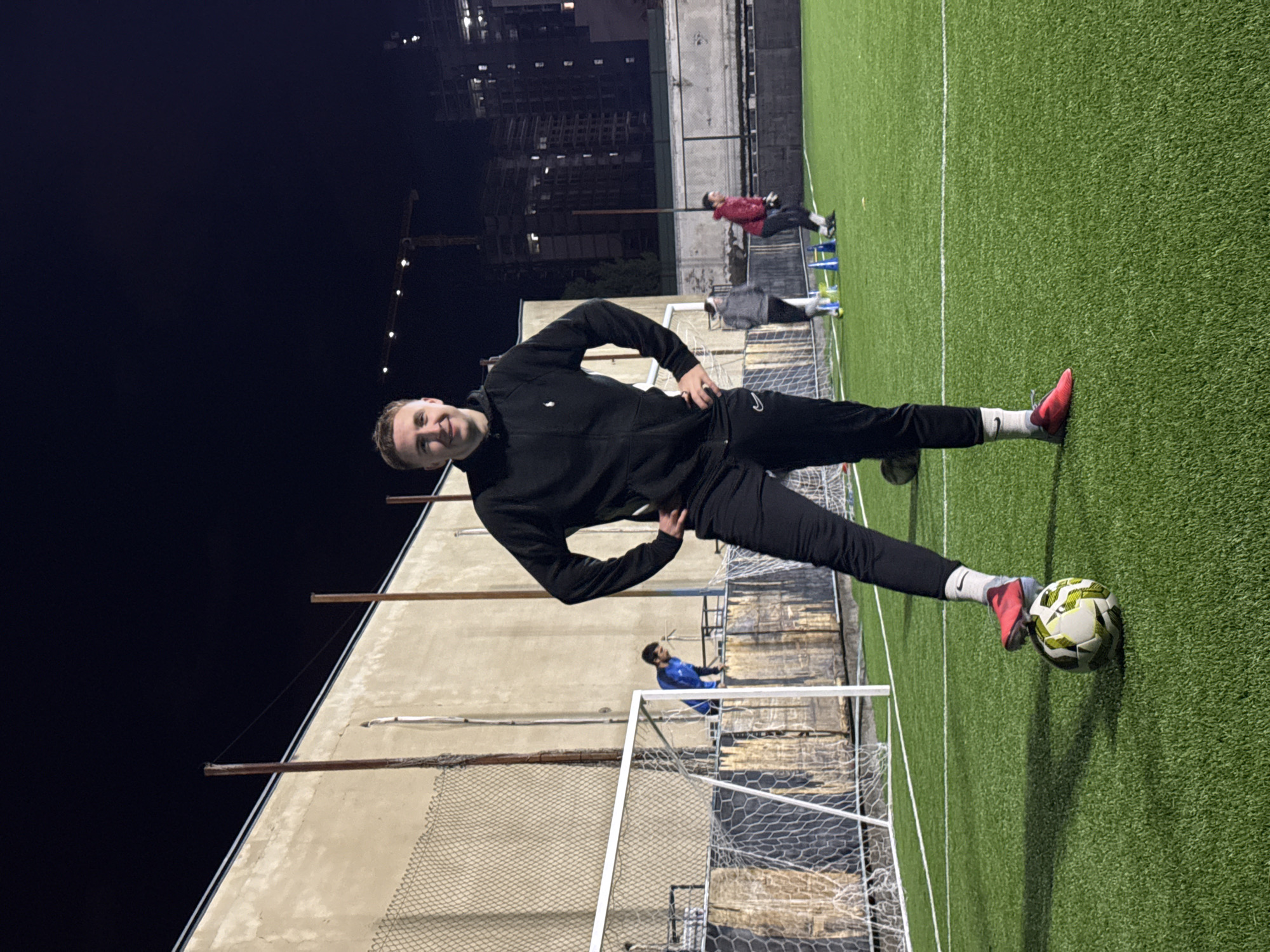 A photograph taken at night on a soccer field in Tbilisi, Georgia shows a young man balancing on a soccer ball with his right foot. He is wearing a black tracksuit, white socks, and red shoes. He is smiling at the camera with both hands on his hips. A soccer goal is to his left, while other people are visible on the field in the background, alongside training equipment. The background shows dark sky over a building, a construction crane, and other buildings in the cityscape.
