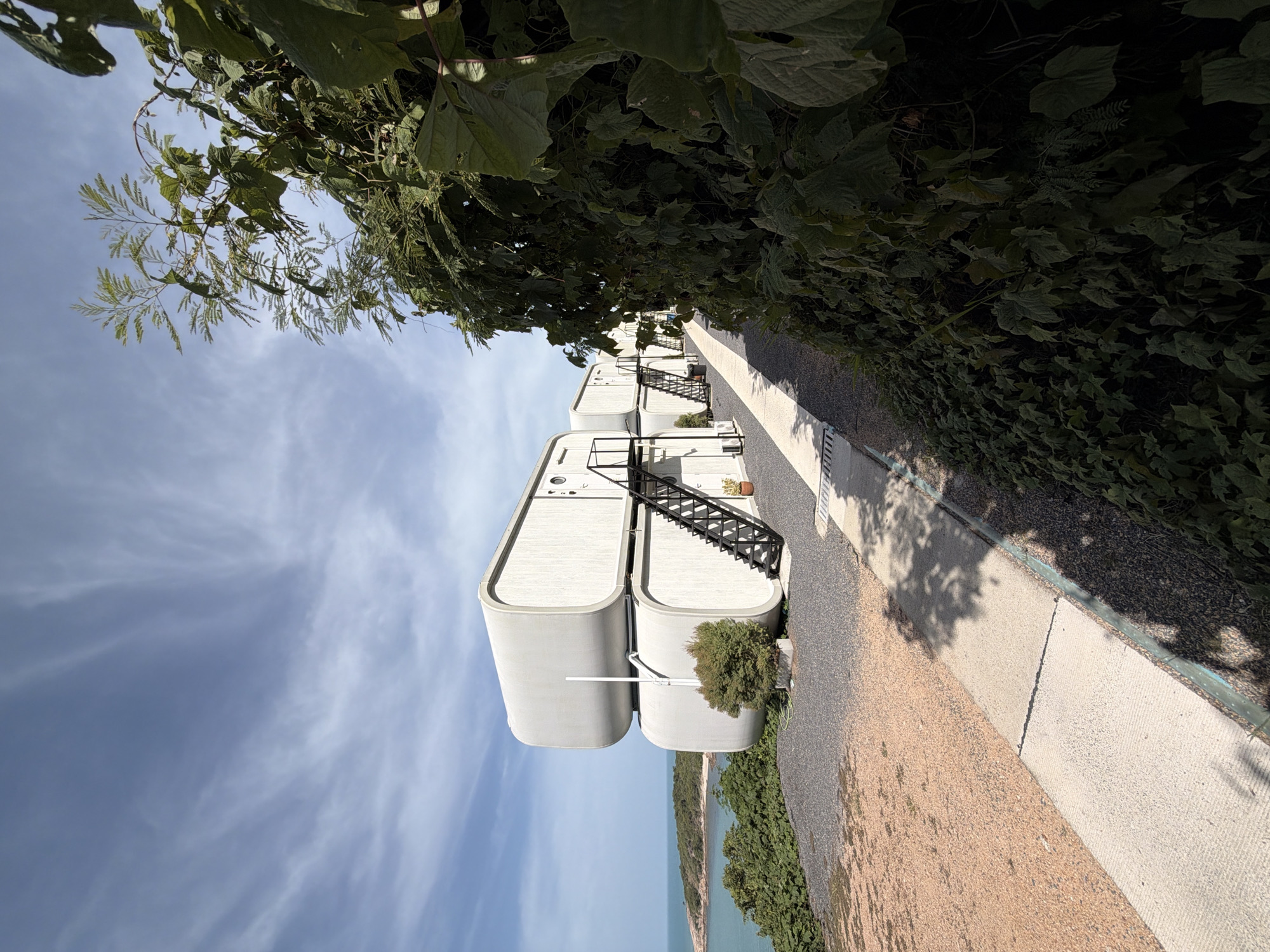 In Ko Samui District, Thailand, a stacked modular hotel unit, characterized by its rounded edges and white exterior, is situated on a hillside overlooking the ocean. A dark metal staircase provides access to the upper unit. The hotel unit is surrounded by a gravel path and terra cotta colored gravel. A paved walkway, bordered by lush green foliage, runs alongside the hotel. In the distance, a body of water and a small island are visible under a bright, cloudy sky. The scene appears tranquil and serene, suggestive of a holiday destination.