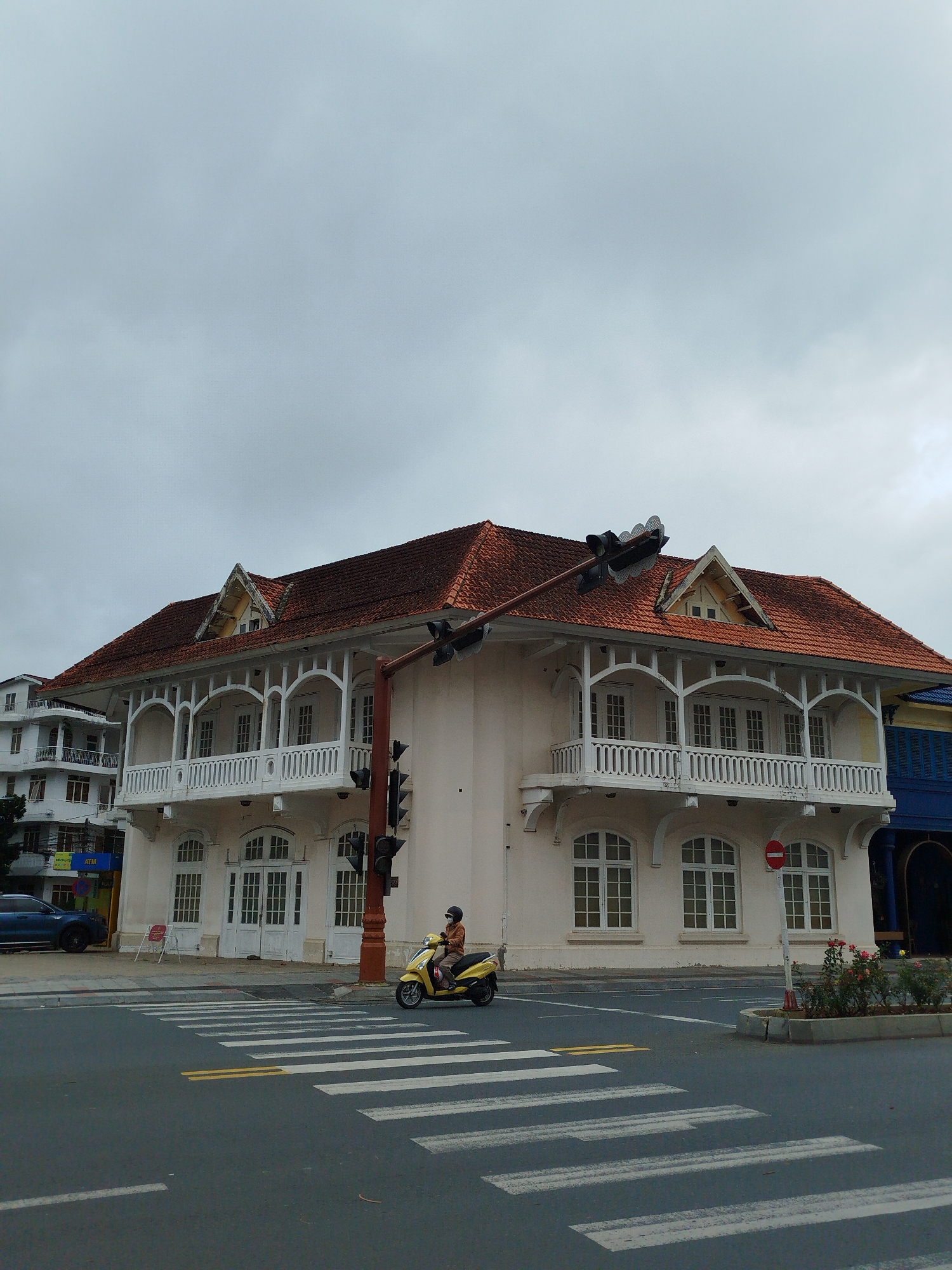 An overcast daytime scene captures a street corner in what appears to be an urban area, with a prominent two-story building dominating the right and center of the frame. The building features a distinctive red tiled roof with multiple gables and dormer-like structures. Its facade is light-colored, possibly cream or white, adorned with an ornate white wooden balcony spanning much of the second floor, supported by decorative arches and columns. The balcony has a detailed balustrade. The ground floor has several arched windows and double doors, also white.
In the foreground, a multi-lane asphalt road with clearly marked white pedestrian crosswalks and yellow double lines leads towards the building. A person wearing a helmet and a face mask is riding a bright yellow scooter, positioned near the crosswalk and a traffic light pole. The traffic light, equipped with multiple signal heads, stands tall next to the building's facade.
To the left, a blue car is parked on the sidewalk next to a smaller, multi-story white building, which also has balconies. A small green plant bed with red flowers is visible on the sidewalk curb to the right of the crosswalk. A red circular sign with a white horizontal bar, indicating "No Entry" or "Do Not Enter," is placed on the sidewalk in front of the main building. A partially visible blue storefront with a corrugated door can be seen to the far right.
The sky is uniformly gray and cloudy, suggesting an overcast day. Visible text includes "ATM" on a blue sign attached to the building on the far left. The overall scene depicts a functional street environment with distinctive architecture.