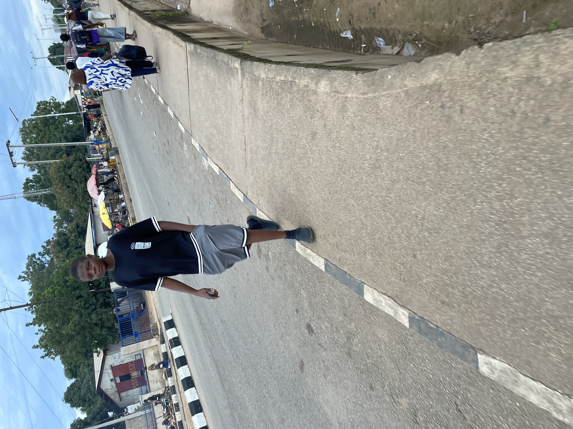 The image depicts a street scene in Jos, Nigeria, under a cloudy sky. In the foreground, a young male is walking along the paved sidewalk, wearing a black t-shirt with white stripes on the collar and sleeves, and grey shorts with white striped trim. He has his right hand in his pocket. The sidewalk runs alongside a paved road with a black and white painted curb. Further down the road, on the opposite side, a market is set up with various stalls and people. Several individuals are visible, some walking and others engaged in activities at the market. Buildings line the street in the background, including what appears to be a small store with a red-painted door. The overall environment suggests an active urban area with commercial activity.