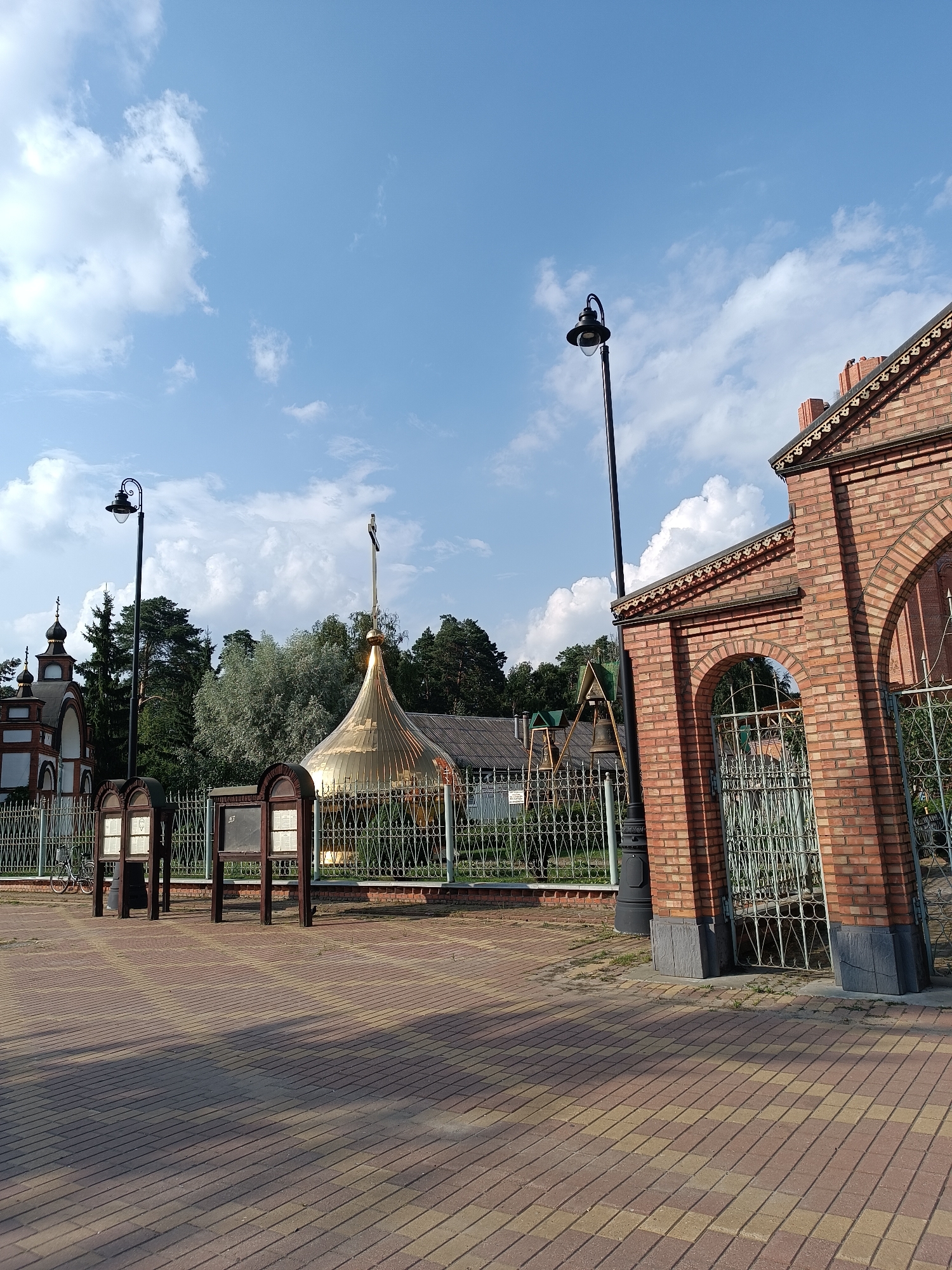 The image depicts a bright, sunny day in Il'inskiy, Russia, featuring a religious complex and its surroundings. In the foreground, a paved walkway made of red and yellow bricks leads the eye towards the structures. To the right, a prominent brick building with arched entrances and a decorative roofline stands, partially obscuring a metal gate. A tall black lamppost stands beside this building.
Behind the walkway and a decorative metal fence, a golden dome with a cross tops a building, suggesting a church or chapel. To the left of the dome, another smaller tower with a dark roof and a cross is visible, also part of the religious complex. The background is filled with lush green trees, indicating a natural, possibly park-like setting. Two wooden information stands are positioned in front of the fence, each displaying notices within glass cases. A bicycle is parked to the left of these stands. The sky is a clear blue, punctuated by fluffy white clouds, contributing to the overall pleasant atmosphere of the scene.