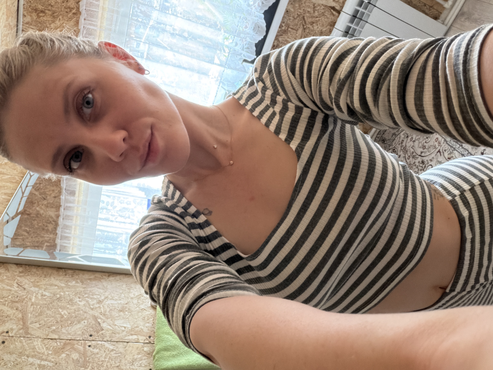 A woman with fair skin and blonde hair is facing the camera, wearing a striped grey and white long-sleeved top. She has blue eyes, and a delicate gold necklace is visible around her neck. The setting appears to be indoors, with a window in the background allowing natural light. The walls appear to be made of wooden boards. The exact location in the city of Chaikovsky, Russia cannot be determined from the image.