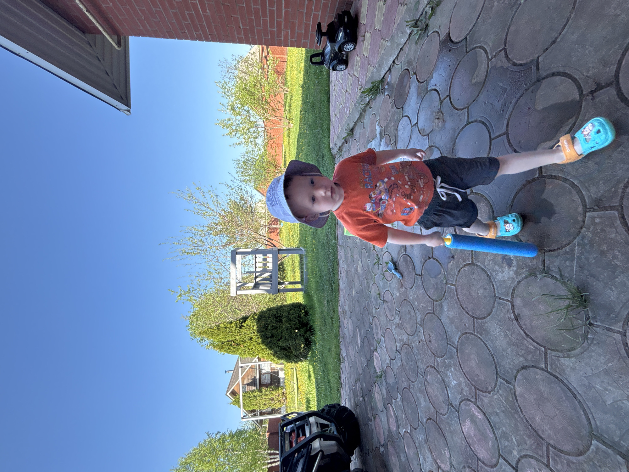 A child stands on a patterned paving stone surface next to a brick building, holding a blue foam object. The child wears a light-colored sun hat, an orange t-shirt, dark shorts, and light blue sandals with orange straps. A black toy car sits near the brick building. A green lawn is visible beyond the paving stones, with trees and a small structure visible in the background. A black all-terrain vehicle is partially visible in the lower left corner. The location appears to be Ферма, Russia.