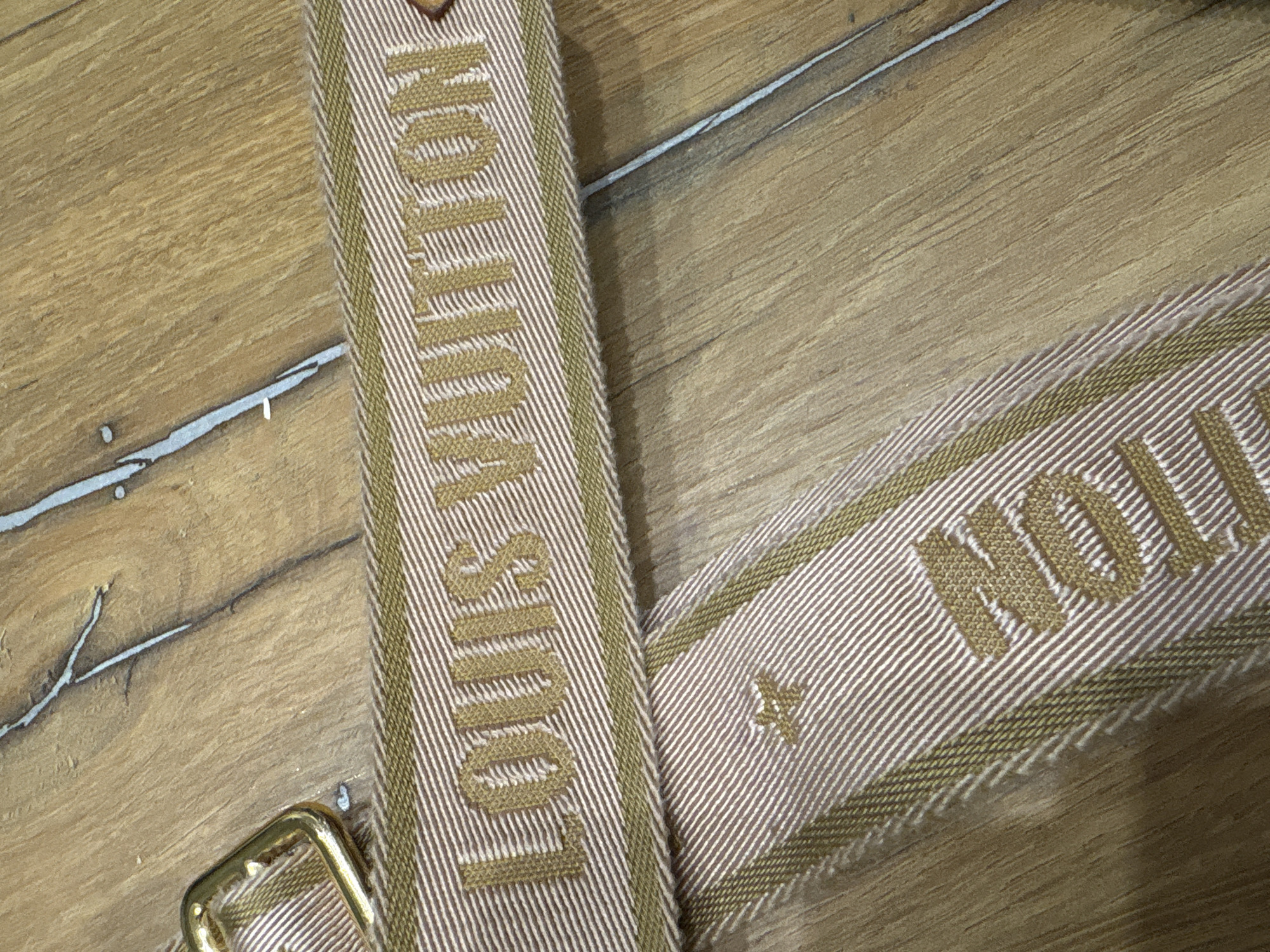 The image shows a close-up of a fabric strap with the Louis Vuitton logo embroidered on it. The strap is a light brown color with white and olive green stripes. The Louis Vuitton logo is embroidered in a golden color. A golden buckle is visible on the left side of the strap. Another part of the strap with the word "TION" embroidered is visible below the logo. The background is a wooden surface with visible grain and some white lines. The location is Unknown, Unknown.