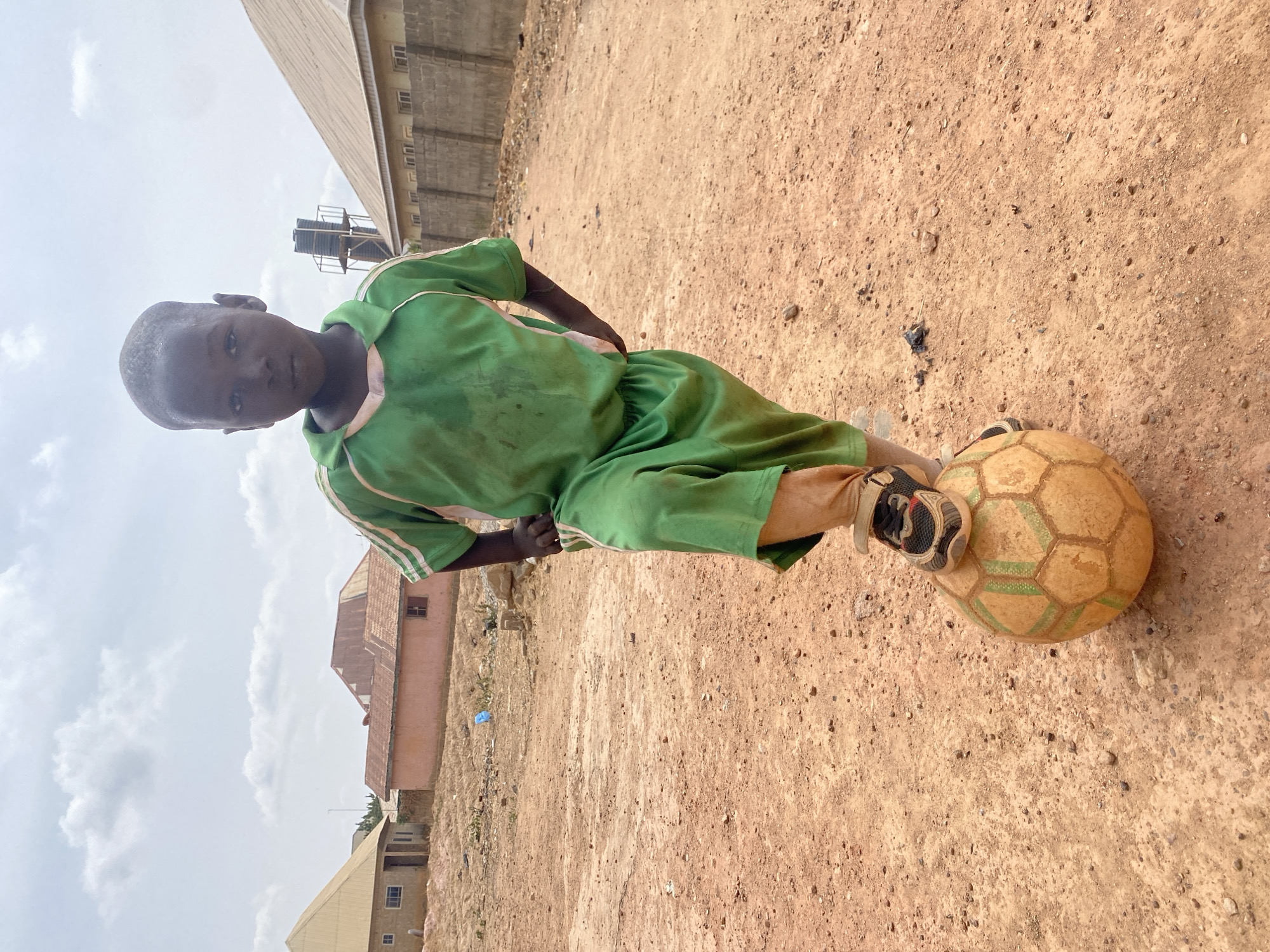 A young male, appearing approximately 6-10 years old, stands centered in an unpaved, reddish-brown dirt area within Rido, Nigeria. He wears a soiled green short-sleeved shirt with white shoulder stripes and matching green shorts, complemented by athletic shoes and light-colored socks. His gaze is directed forward. His right foot rests on a worn, tan and green hexagonal-patterned soccer ball positioned on the ground. His hands are placed on his hips. The background consists of multiple structures under a partly cloudy sky. To the left, a light-colored building with a yellow corrugated roof and a reddish-pink building with a red corrugated roof are visible. To the right, a light-colored building with a sloped roof is partially obscured by a grey concrete block wall. A tall metal structure supporting two dark water tanks is present in the distant right background.