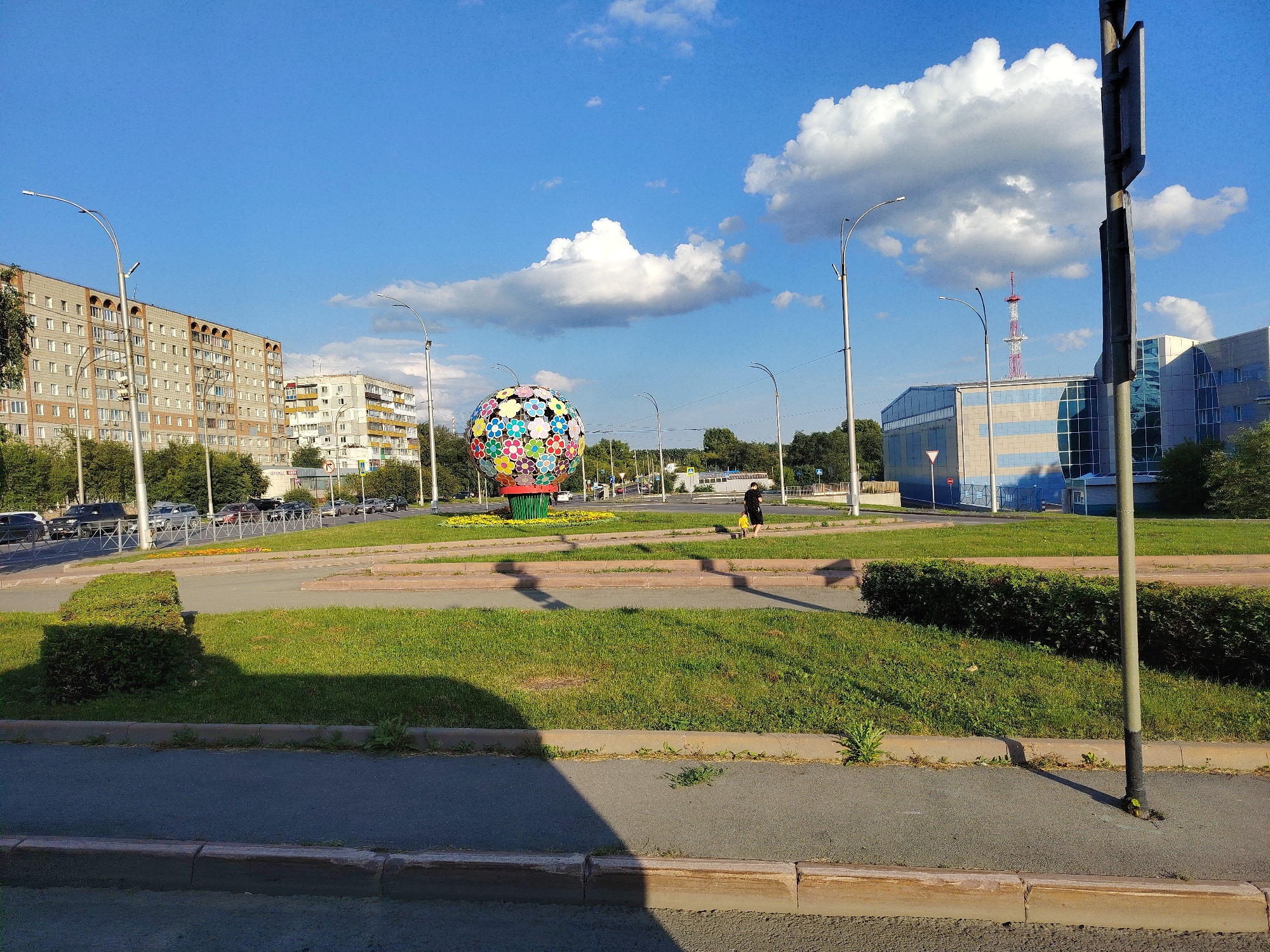 The image captures a street scene in Kemerovo, Russia, under a clear blue sky with scattered white clouds. A large, spherical sculpture decorated with colorful flowers is prominently featured in the center of a roundabout. Tall apartment buildings, typical of Soviet-era architecture, line the left side of the frame, while a modern glass-fronted building stands on the right. A communication tower with a red and white antenna is visible in the background. The street is busy with traffic, and a person in a yellow shirt and black pants walks on the sidewalk. The foreground shows a paved road with a grassy median and hedges. The overall impression is one of an urban environment with public art and everyday city life.