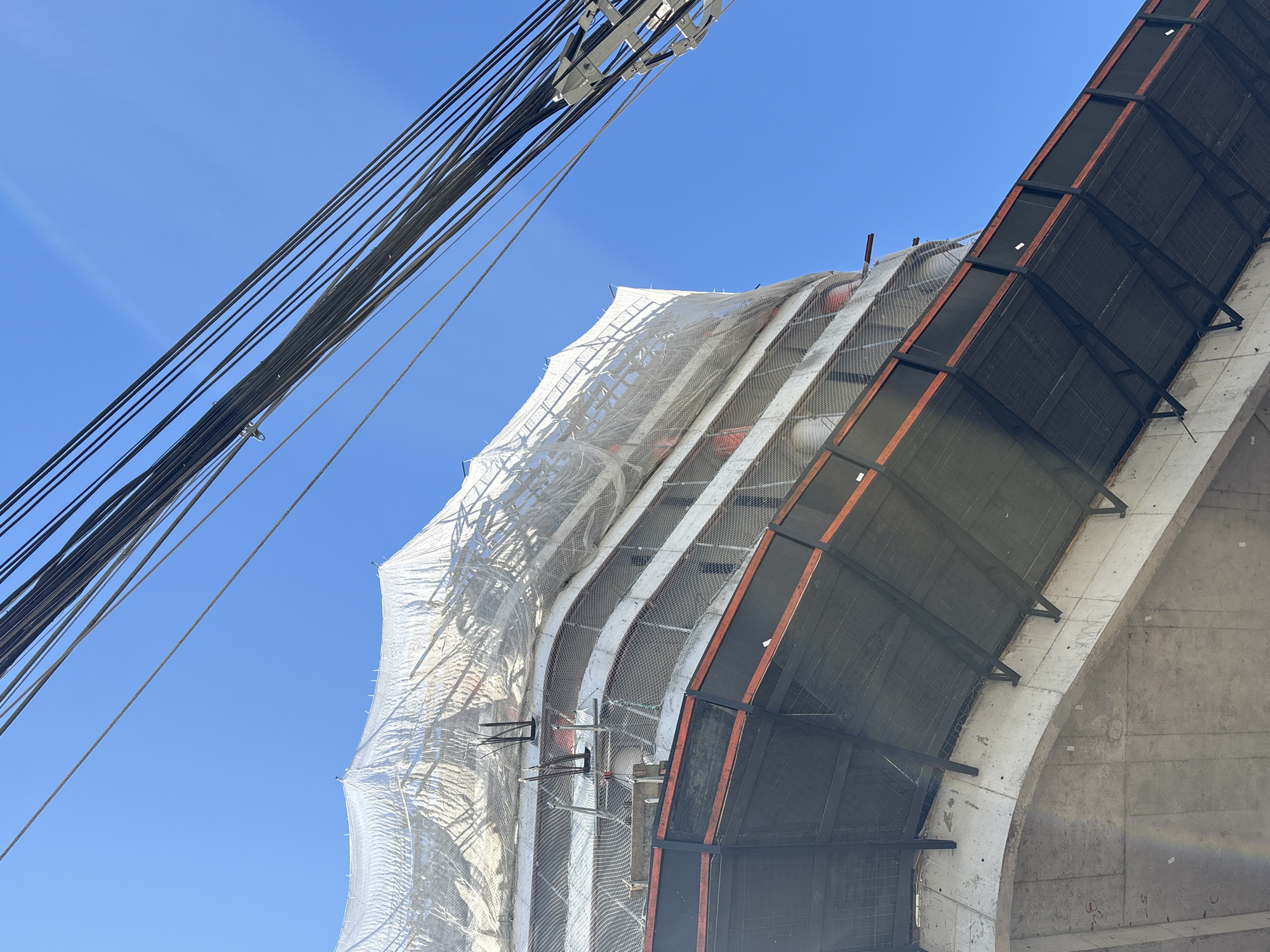 The image shows a construction site with a tall building under construction, covered in a white protective tarp. The building has a curved facade and appears to be made of concrete. There are metal scaffolding and safety nets visible. Overhead, several power lines are strung across the frame, crossing a bright blue sky. The angle of the shot suggests the photo was taken from a lower vantage point, looking up at the construction. The specific location in São Paulo, Brazil, cannot be determined from the image alone.
