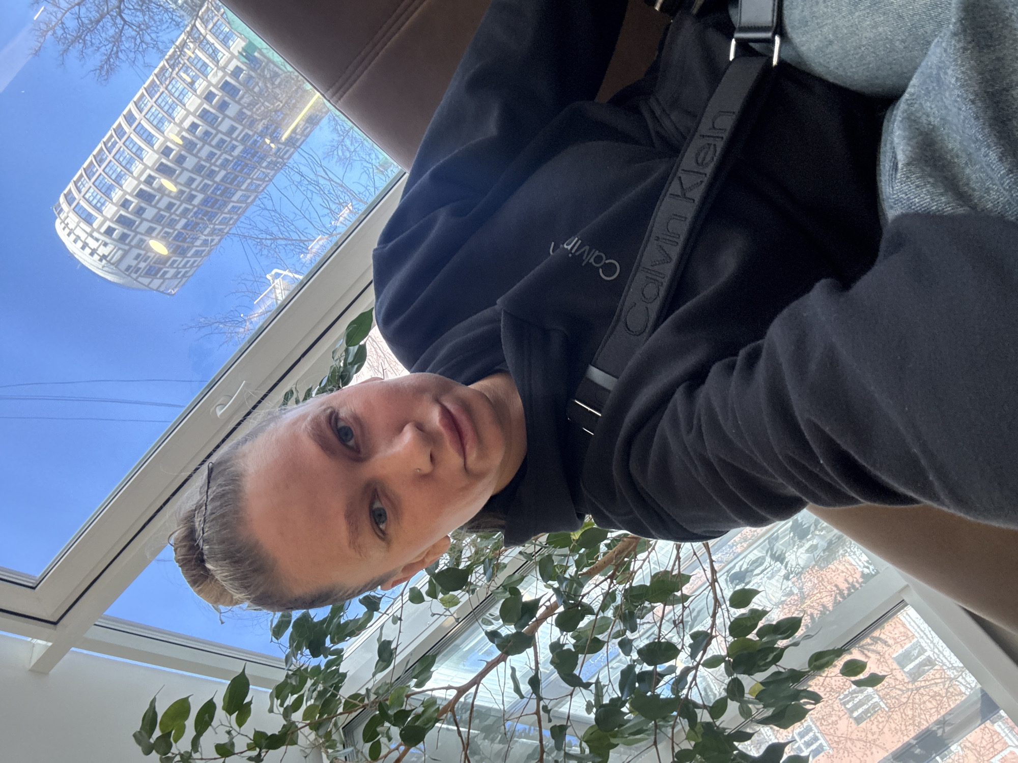 A selfie of a fair-skinned person with blonde hair, wearing a black Calvin Klein hoodie and a shoulder strap with "Calvin Klein" printed on it, sitting indoors with a plant nearby. Through the window, a curved building is reflected in the glass. Outside, the weather is sunny with a blue sky. Based on the architecture visible in the window, the location is Perm, Russia.