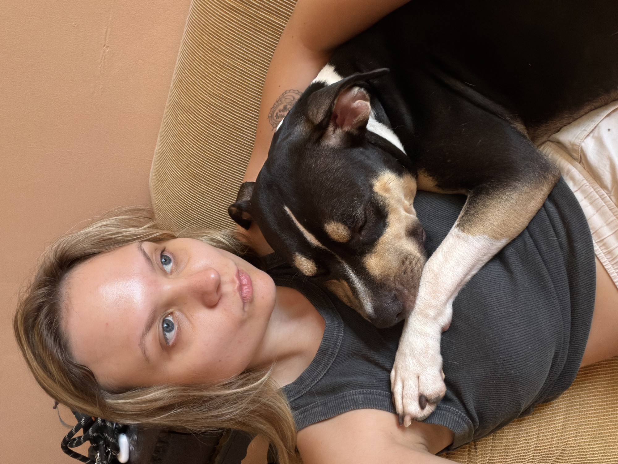 A blonde woman with blue eyes is holding a black and tan dog on a couch. The dog is resting its head on the woman's chest and its paw is draped over her arm. The woman is wearing a dark grey tank top and light pink shorts. She has a tattoo on her right arm. The couch is a golden yellow color with a ribbed texture. The wall behind the couch is a peachy orange color. The scene appears to be indoors, likely in a residential setting in Tel Aviv, Israel.