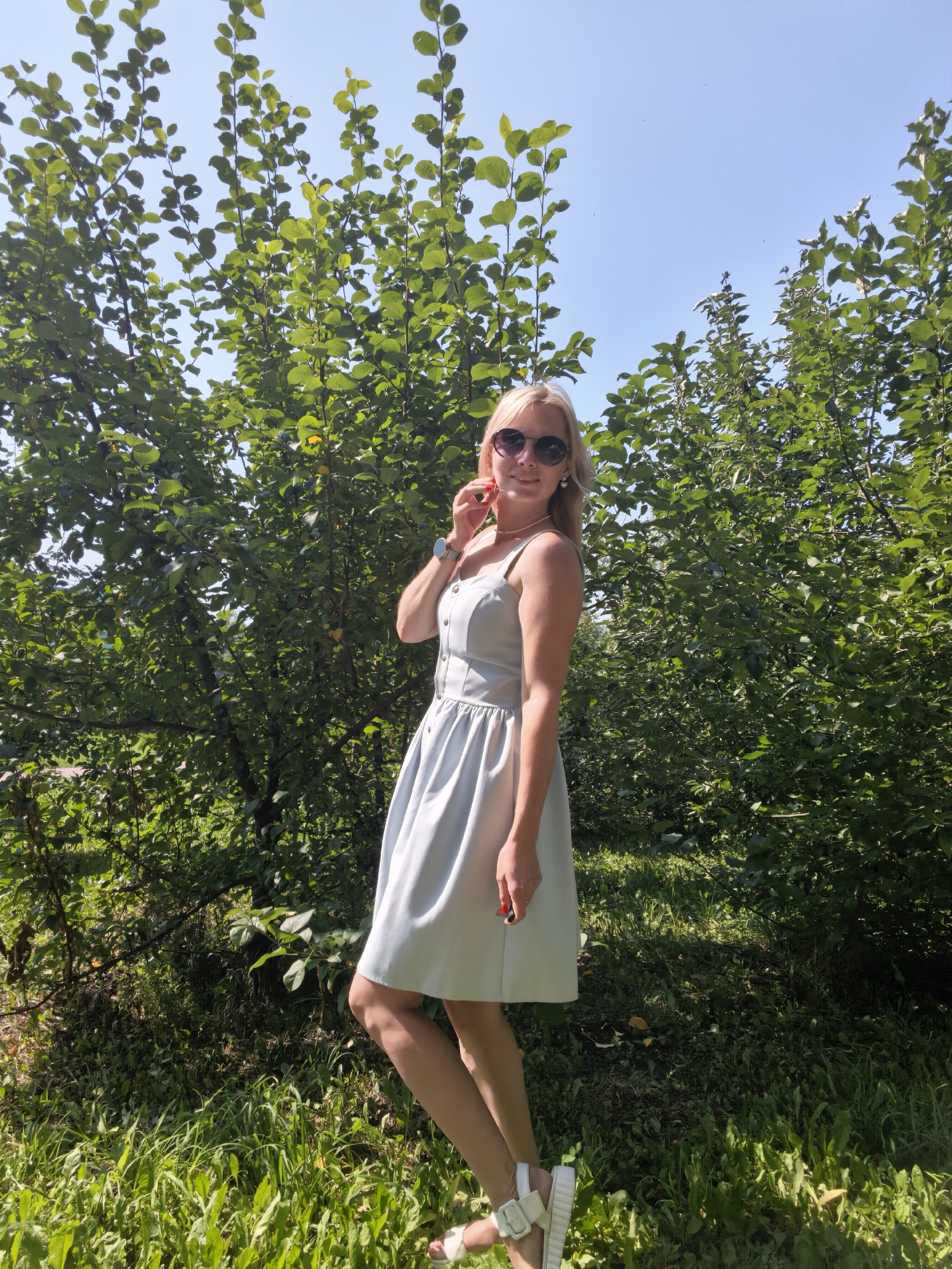 A blonde woman with sunglasses and a light blue dress stands in front of lush green trees and a blue sky. The woman is holding her hand up to her face, and she is wearing white sandals. The image appears to be taken outdoors, possibly in a park or garden, with sunlight illuminating the scene. The location is identified as Sterlitamak, Russia.