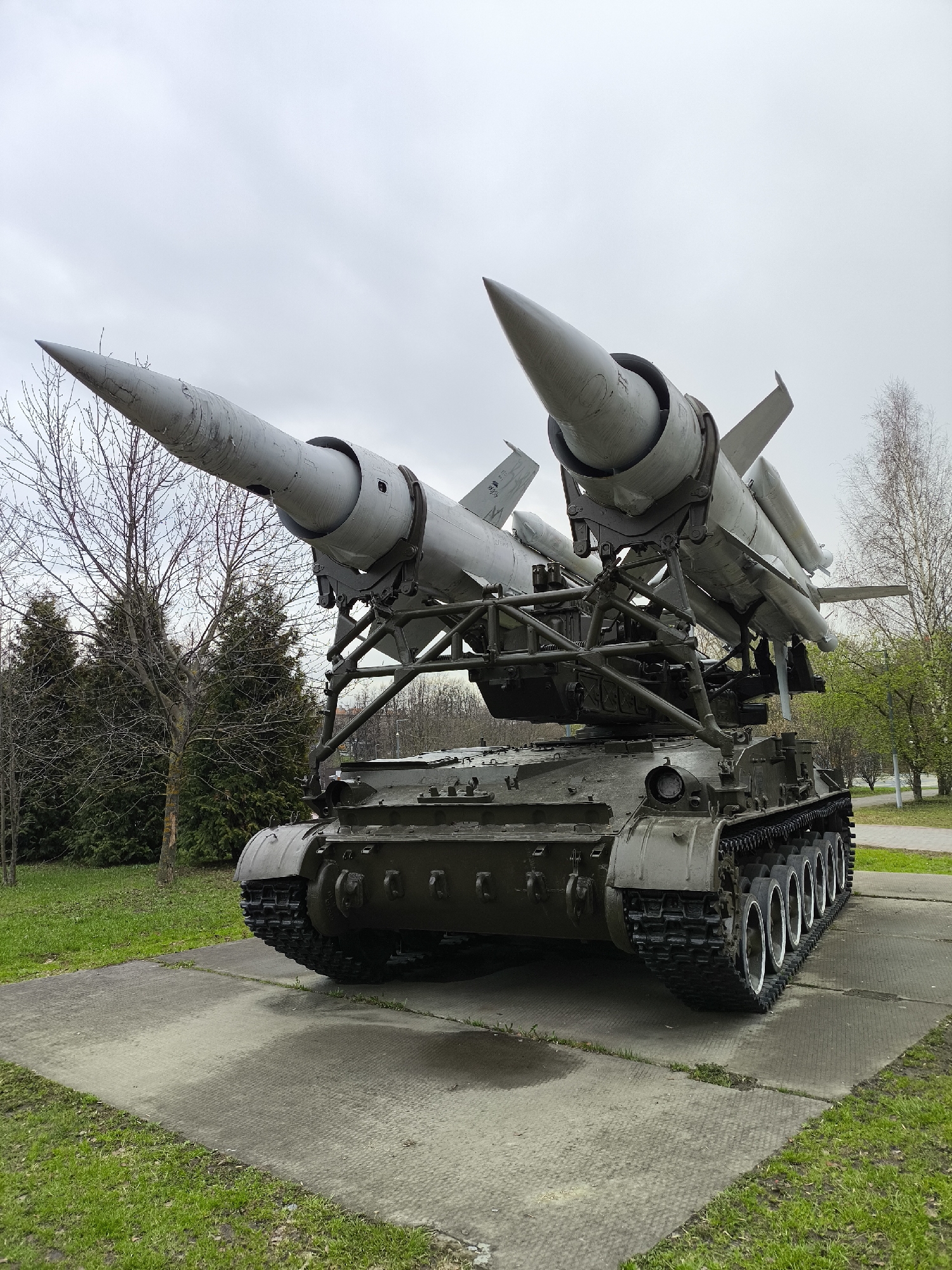 A large, self-propelled anti-aircraft missile system is the dominant subject of the image, displayed outdoors on a concrete slab. The military vehicle features a dark green, tracked chassis, similar to that of a tank, with prominent road wheels visible along its side. Mounted atop the chassis is a launch platform, holding four large, gray, finned missiles. Two missiles are positioned prominently in the foreground, angled upwards and outwards, showcasing their pointed noses and weathered surfaces. Two additional missiles are partially visible behind them.
The system is set within a park-like environment. Surrounding the concrete display area is vibrant green grass. The background consists of a mix of trees: bare-branched deciduous trees, suggesting late winter or early spring, are interspersed with darker green evergreen trees to the left. On the right, deciduous trees appear to be just beginning to bud with new green leaves.
The sky is uniformly overcast and light gray, indicating a cloudy daytime setting. The concrete slab beneath the vehicle shows some damp patches, possibly from recent rain. No people or active events are depicted; the missile system appears to be a static exhibit.
Visible, albeit faint, text can be seen on the tail fins of the missiles. On the left missile's fin, numbers resembling "9 47" or "9 7" are partially discernible. On the right missile's fin, blurry characters that might be "M4" or "M9" are present.