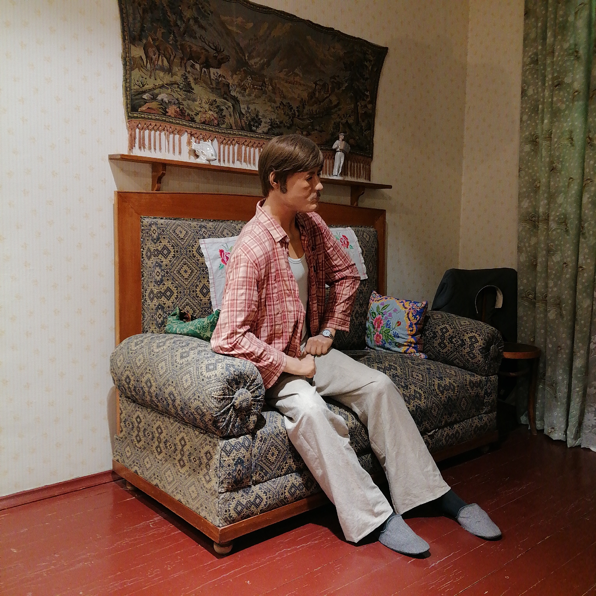 A wax figure of a middle-aged man is sitting on a sofa in a room. The man has brown hair and a mustache. He is wearing a pink plaid shirt, a white tank top, beige pants, and gray slippers. He has a watch on his left wrist. The sofa has a patterned upholstery and several pillows. Behind the sofa is a wooden headboard with a shelf. On the shelf are two decorative figurines. Above the headboard hangs a tapestry depicting a forest scene. To the right of the sofa is a small wooden table with a black object on it. A patterned curtain hangs in the background on the right side of the image. The walls are covered in patterned wallpaper. The floor is red painted wood. The location is Moscow, Russia.