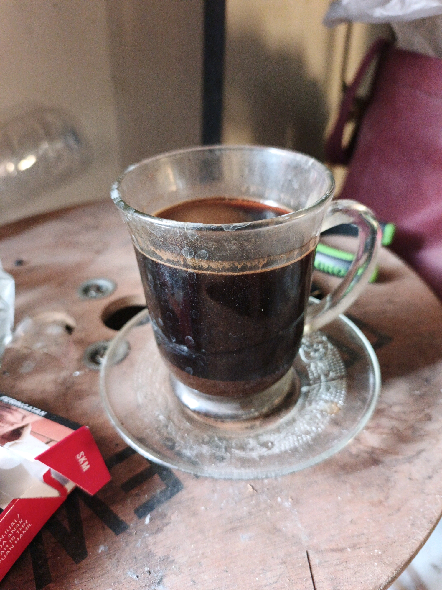 A glass cup of coffee sits on a saucer on a wooden surface. The coffee is dark brown and appears to be freshly brewed. The cup and saucer are made of clear glass and have a simple design. The wooden surface is a rough, textured wood, possibly a table or a spool. In the background, there are blurry shapes of objects, including what looks like a red bag and some plastic bottles. To the left of the cup, there is an open red and white cigarette pack. The scene is lit by natural light, creating soft shadows. The image appears to be taken indoors, possibly in a home or a casual establishment. The presence of the cigarette pack suggests a setting where smoking is permitted. The overall impression is one of a simple, everyday moment, perhaps a break or a moment of reflection. It is not possible to determine the specific location in Ketegan, Indonesia based solely on this image, as there are no identifiable landmarks or contextual clues.