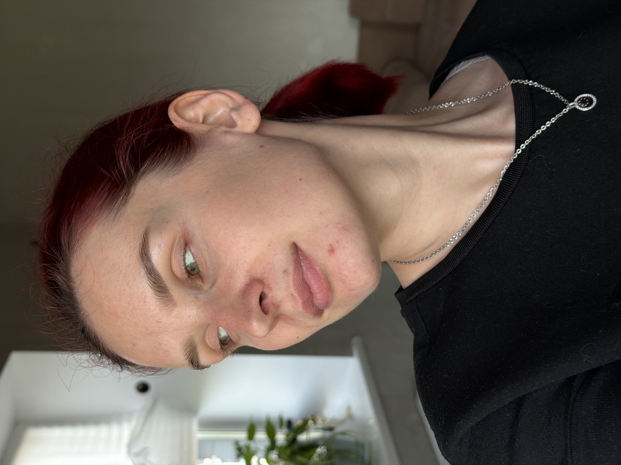 A woman with red hair pulled back into a ponytail is depicted in a close-up, sideways-oriented photograph. She is wearing a black top and a delicate silver necklace with a small pendant. Her skin is fair with some visible blemishes, and her expression is pensive. The background is indistinct, showing a muted beige wall and a blurred view of a window and indoor plants. There is no contextual information to indicate the location is Chaikovsky, Russia.