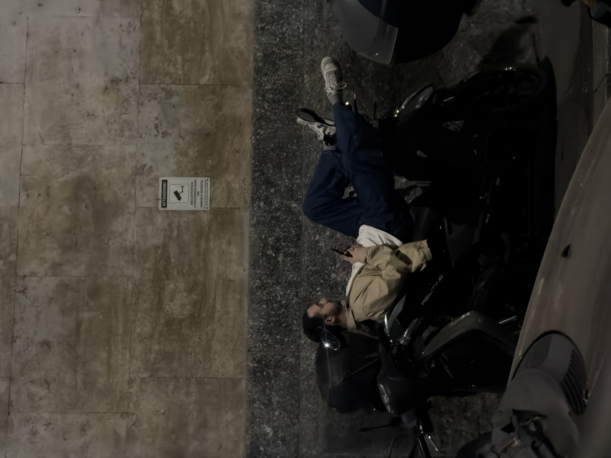 A person lies atop a scooter on a street next to a light-colored stone wall. The person is wearing jeans, sneakers, a light-colored shirt, and a tan jacket. The person is holding a cell phone. Next to the wall is a sign showing a surveillance camera. A car is on the right side of the scooter. The scene is on a street at night in Milan, Italy.