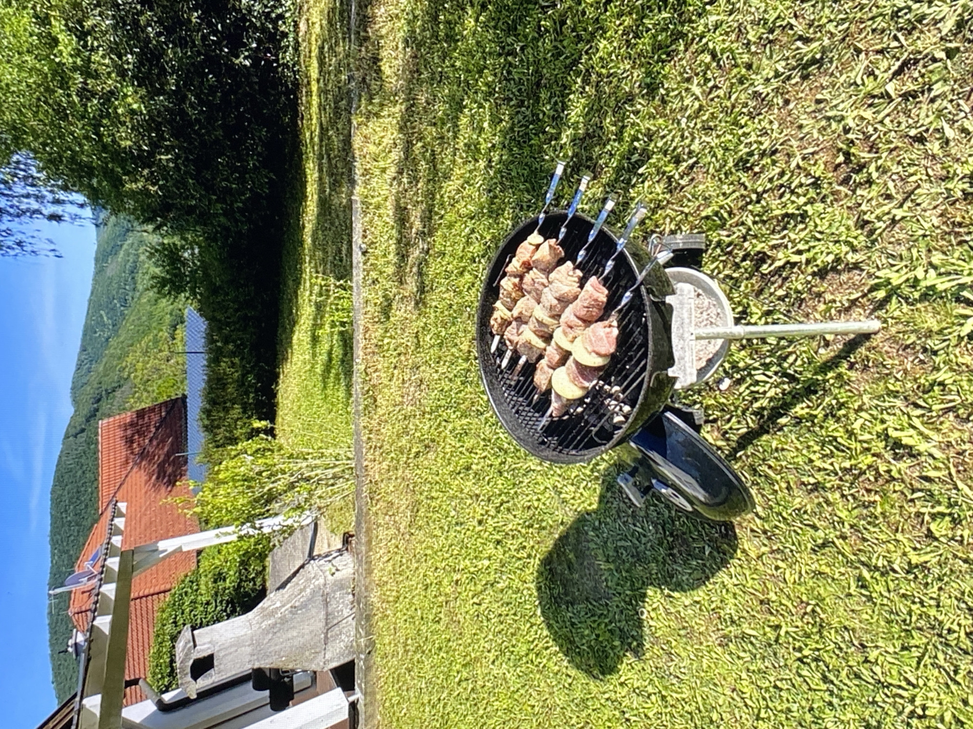 The image depicts a backyard barbecue scene on a sunny day, likely in Purkersdorf, Austria, given the architectural style of the house and the surrounding greenery. In the foreground, a black kettle-style charcoal grill is positioned on a metal stand, laden with skewers of meat and what appear to be slices of potato. The grill is on a well-maintained green lawn, casting a distinct shadow to the left. Behind the grill, a portion of a covered outdoor seating area or veranda is visible, with white support beams and a grey stone chimney structure. Further in the background, a red-tiled roof house with white trim is partially visible, nestled amongst lush green trees. Rolling hills covered in dense foliage rise behind the house, under a clear blue sky. The overall impression is one of a peaceful, domestic outdoor setting.