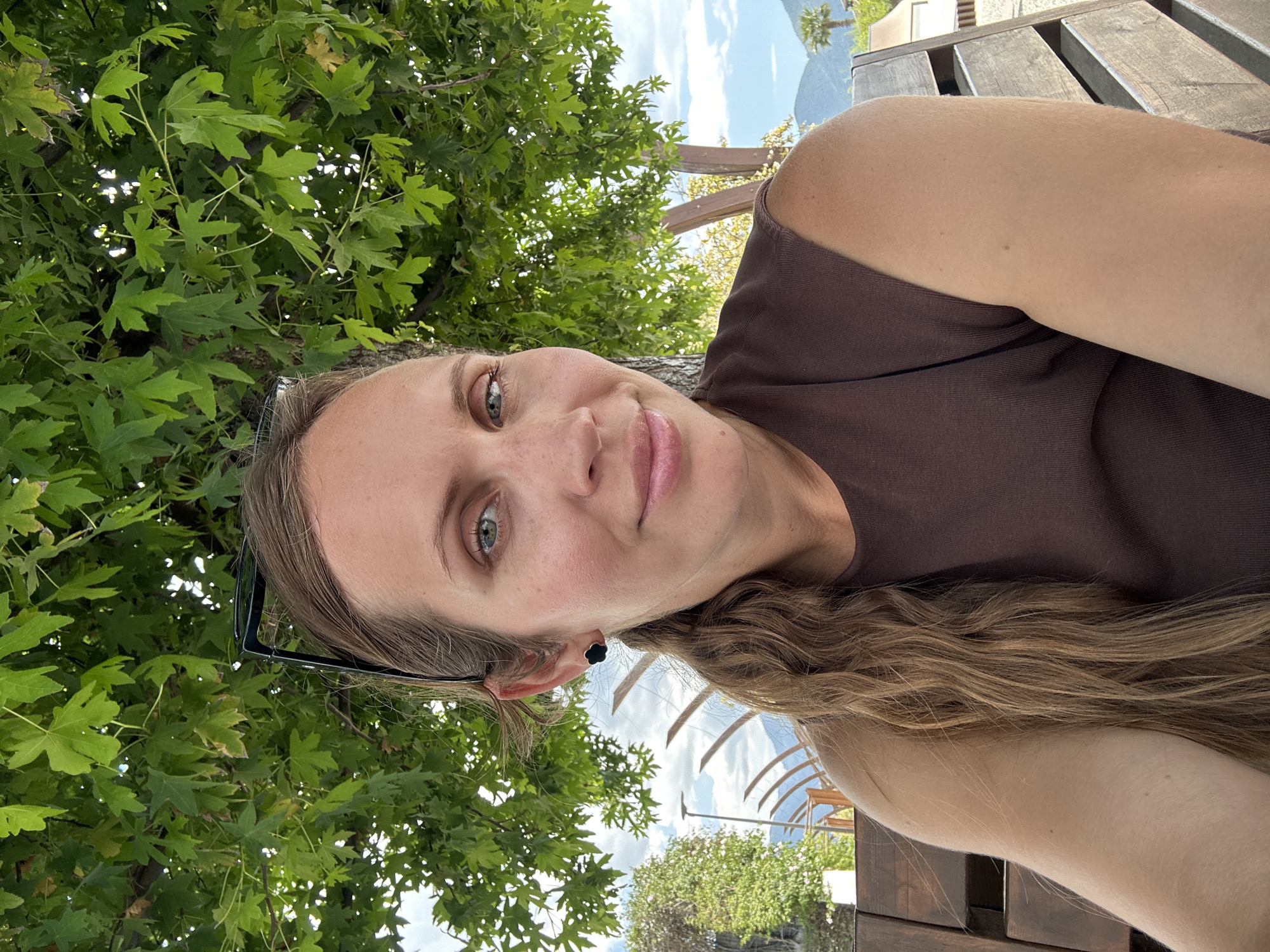 A medium shot depicts a woman with long, wavy brown hair, wearing a dark brown sleeveless top, and sunglasses are tucked into her hair, resting behind her left ear. She has fair skin, light eyes, and a slight smile. She is outside, with lush green foliage filling much of the background. In the upper part of the frame, between the leaves, is a view of a blue sky with scattered clouds, and what seems to be a body of water with a distant mountain or island. The woman appears to be seated on wooden slats. There is partial view of an architectural structure or pergola in the background. The city of Fethiye, Turkey is not discernible in the image.