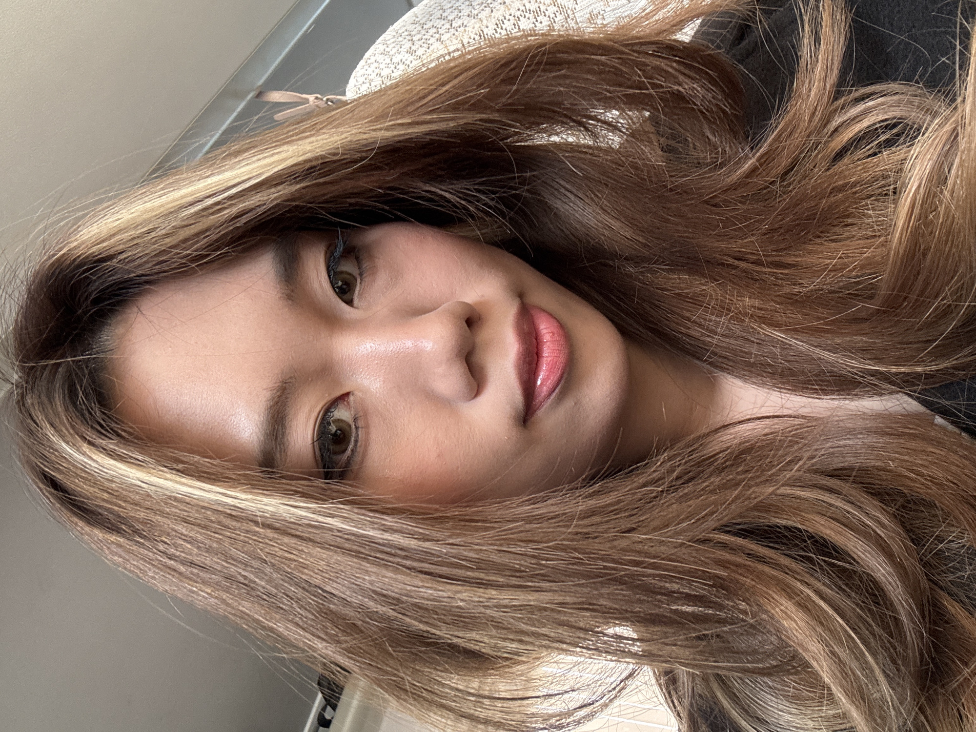 A close-up, portrait-style image of a young woman with light brown hair with blonde highlights is presented. Her skin is fair with a warm undertone, and she has brown eyes. She is wearing makeup, including eyeliner, blush, and lip gloss. The background is out of focus, but parts of a light-colored object, possibly a hat, are visible along with a hint of a wall. There is no visual information in the image to suggest a specific location in Guazhou County, China, or any other geographical location. The image focuses solely on the subject's face and hair.