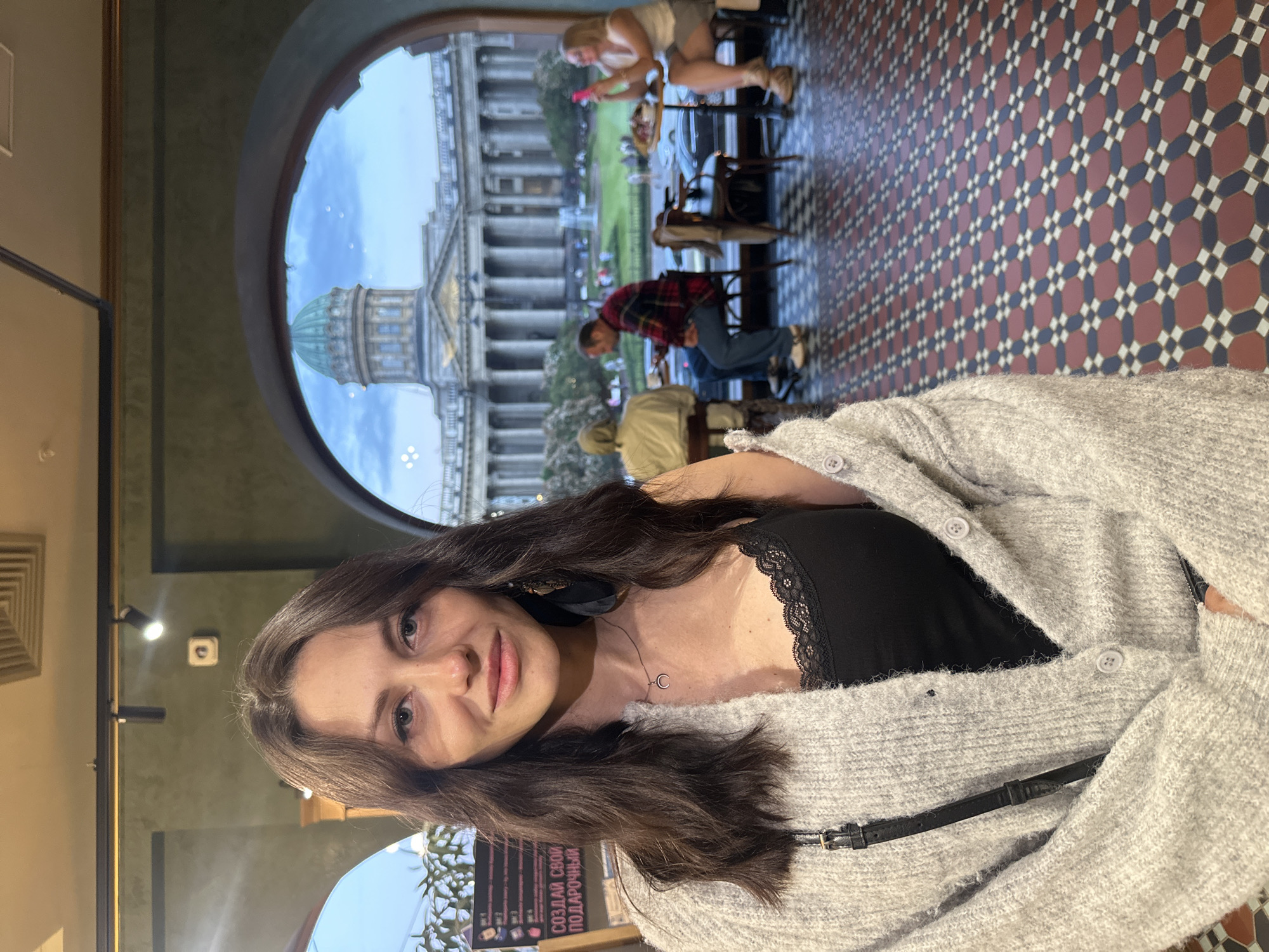 A medium shot shows a woman with long, dark, wavy hair and fair skin standing indoors, facing the camera. She is wearing a gray, fuzzy cardigan over a black lace-trimmed tank top and a silver necklace with a crescent moon pendant. A black strap of a bag is visible across her chest.
In the background, through an arched opening, a scenic view of Kazan Cathedral in Saint Petersburg, Russia, is visible. The cathedral features a prominent green dome and a colonnade. In the outdoor area beyond the cathedral, several people are seated at tables outside a building, and a fountain is visible in the distance. The floor inside the building is covered with a patterned tile mosaic of red, black, and white geometric shapes. The overall lighting suggests it is daytime.