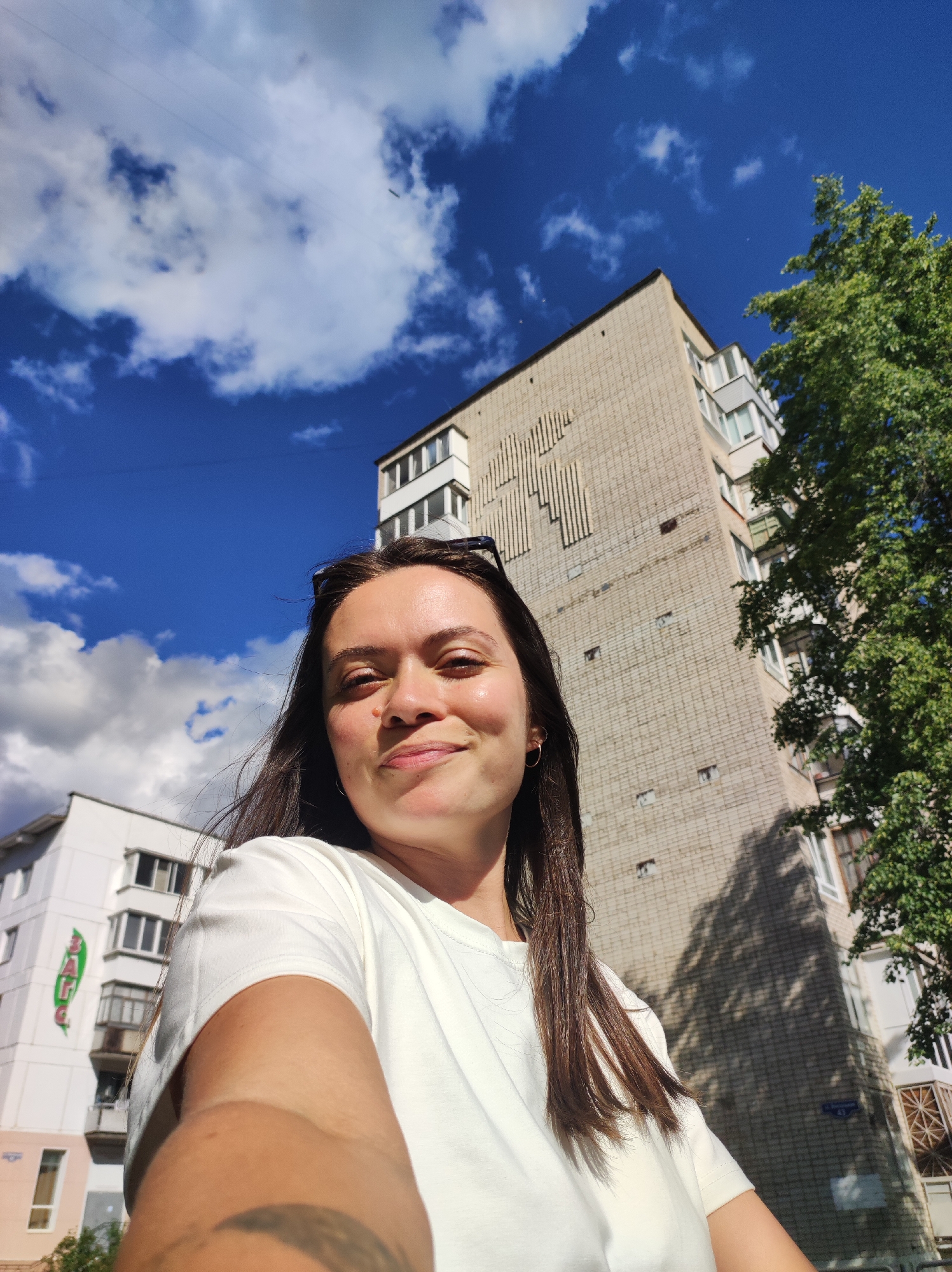 A selfie of a woman with long brown hair and wearing a white t-shirt. Sunglasses are perched on her head. She is smiling slightly. In the background is a clear blue sky with white puffy clouds, a large concrete apartment building with a decorative design, and a green tree. In the lower left corner, a partially visible white building is also present. Based on the details visible, the location is likely in the city of Chaikovsky, Russia.