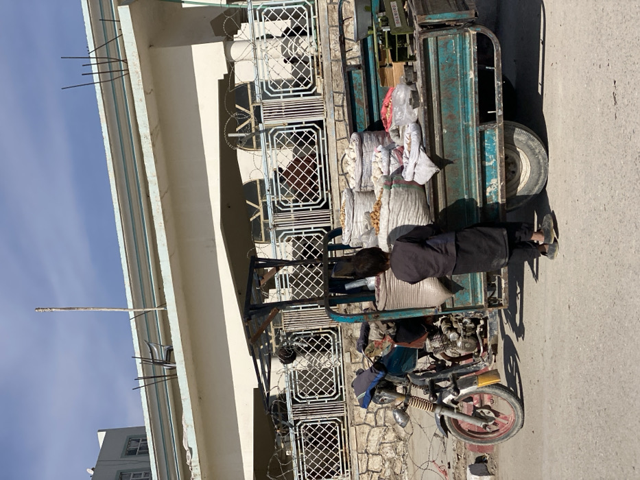 The image depicts a street scene in Kandahar, Afghanistan, under bright daylight. A young child, viewed from behind, wearing a dark long-sleeved shirt and dark trousers, stands on a paved road facing a stationary, teal-colored three-wheeled motor-rickshaw. The vehicle's cargo bed is loaded with multiple large, white sacks containing what appear to be potatoes and other produce, along with smaller bags. A green "DEA WING" weighing scale is positioned on the right side of the cargo bed. A dark-clad individual is partially visible within the motor-rickshaw's driver area.
In the background, a light-colored, multi-story building features a balcony or terrace with a white decorative metal railing. Multiple strands of concertina wire are prominently strung along the top of this railing. Arched windows are visible behind the security wire and railing. Below the railing, a stone wall with a concrete cap forms part of the building's structure. The flat roof of the building exhibits exposed rebar, a vertical wooden pole, and a dark chair. The sky is clear blue with minimal clouds. Shadows present on the road and vehicle indicate direct sunlight.
