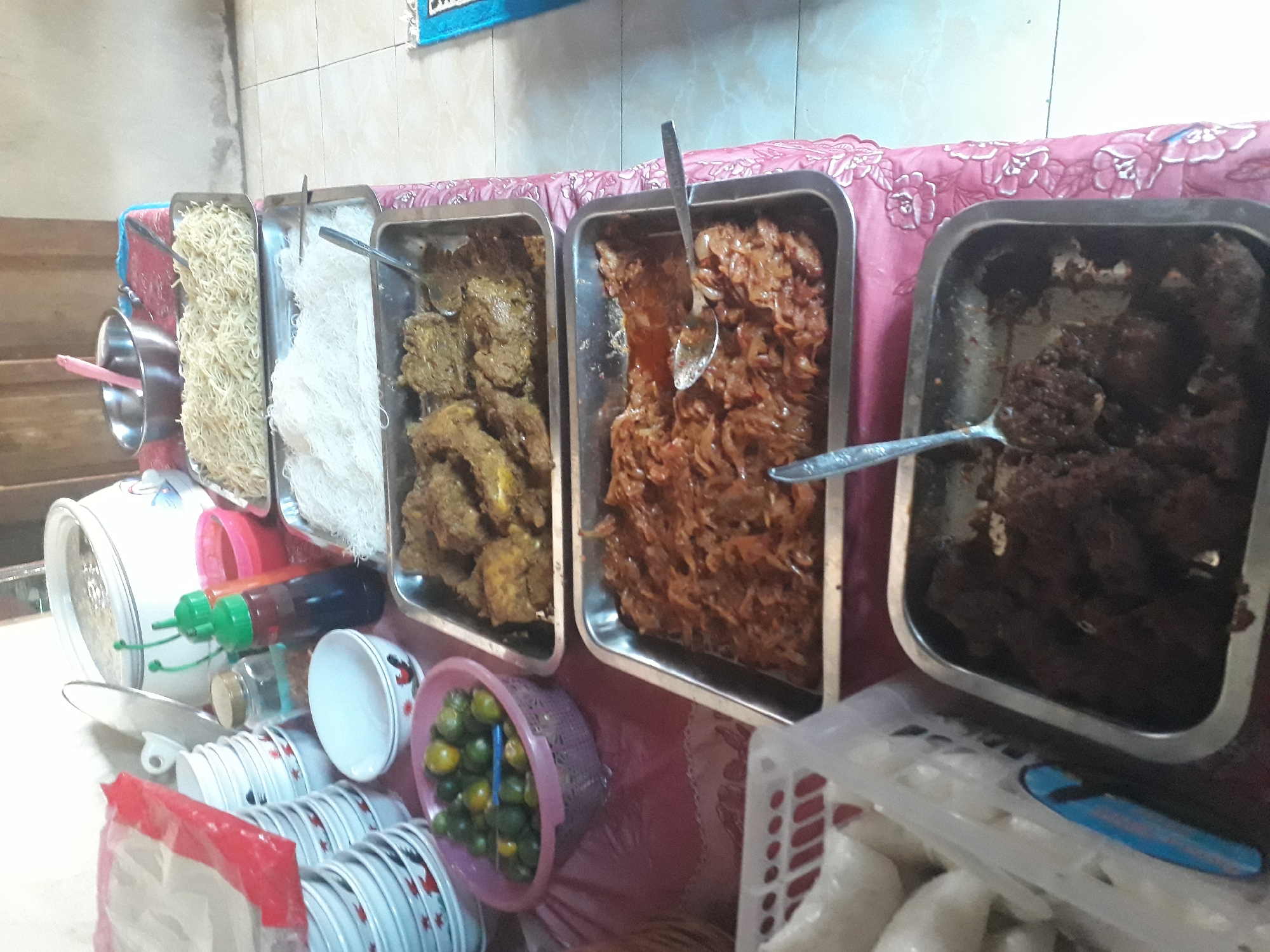 In Desa Balun Ijuk, Indonesia, a table is laden with various dishes, suggesting a meal or gathering. The scene is indoors, with a pale wall and tiled floor visible. The table is covered with a pink tablecloth featuring floral patterns.

Several trays hold food: one contains pieces of what appears to be fried meat or chicken in a dark sauce. Another tray has shredded meat or vegetables in a reddish-brown sauce. Above these, two trays hold noodles – one with thin, white rice noodles and the other with slightly thicker, yellowish noodles. A pink bowl filled with small, green citrus fruits, likely limes or calamansis, sits to the left, next to a stack of white bowls with red and black patterns. To the right of the bowls, there are two squeeze bottles, one red and one green, containing condiments. A large white pot with a lid and a silvery handle is visible in the background, as is a stainless steel bowl containing a pink spatula. To the far left, a white plastic basket contains several white, rectangular food items wrapped in clear plastic, possibly rice cakes.

The overall impression is a spread of food ready to be served, with a focus on savory dishes and accompaniments. The lighting suggests it might be daytime. There are no people visible in the image.