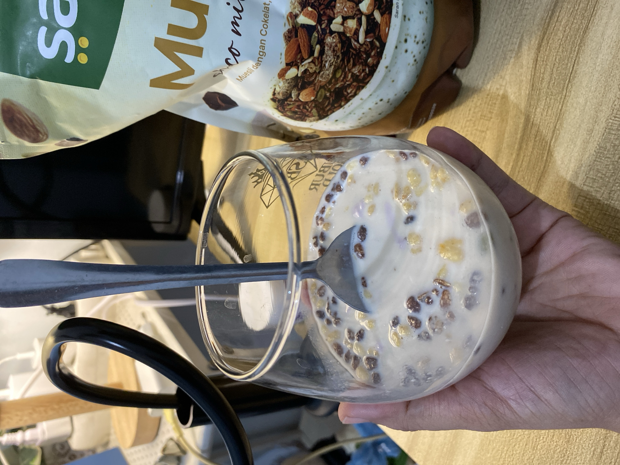 A person's hand is holding a clear glass filled with muesli and milk. A metal spoon is submerged in the muesli. In the background, a bag of muesli is visible, featuring almonds and chocolate on its packaging. The scene appears to be indoors, possibly in a kitchen or dining area, with artificial lighting and a wooden surface beneath the glass and bag. The setting is identified as Babelan, Indonesia.