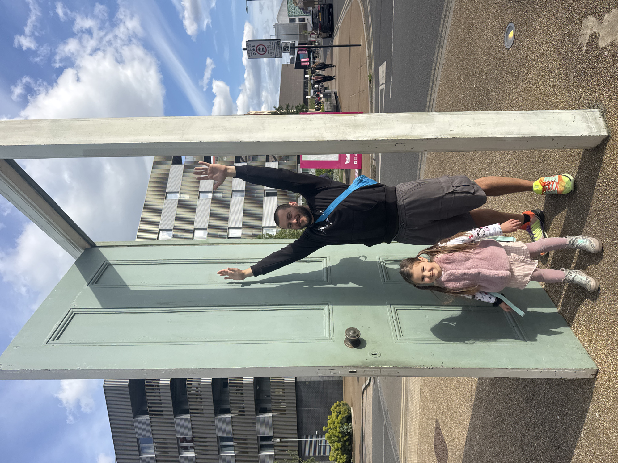 A medium, eye-level shot depicts a man and a young girl posed horizontally, as if they are floating through a large, green doorframe structure. The scene is set outdoors in an urban location.
The man is positioned higher, with his arms extended out to the sides. He wears a black long-sleeved top, grey shorts, and colourful running shoes. A blue bag is slung across his chest.
The young girl is below the man, also with her arms slightly outstretched. She is dressed in a light pink top, a lighter skirt, and patterned leggings with grey boots.
The green doorframe they are posed on is supported by two grey pillars. The background features buildings with various architectural designs. A street with traffic and pedestrians is visible further back. Above, the sky is partly cloudy.
Based on visual landmarks like the pedestrian zone sign and architectural style, this is likely a public art installation in the city of London, United Kingdom.