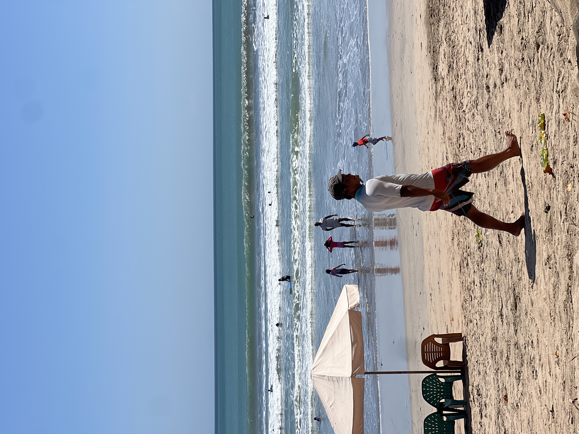 The image depicts a sunny day at Kuta Beach, Indonesia, with a clear blue sky and gentle waves rolling onto the shore. In the foreground, a man wearing a white shirt and patterned swim shorts walks across the sand. To his left, under a large beige beach umbrella, are three plastic chairs, two green and one brown. Further back, in the shallow water, a group of people are visible: two children in swimsuits, a woman in a pink swimsuit, and a man. In the ocean, several individuals are swimming and surfing. The beach is sandy, with some scattered debris.