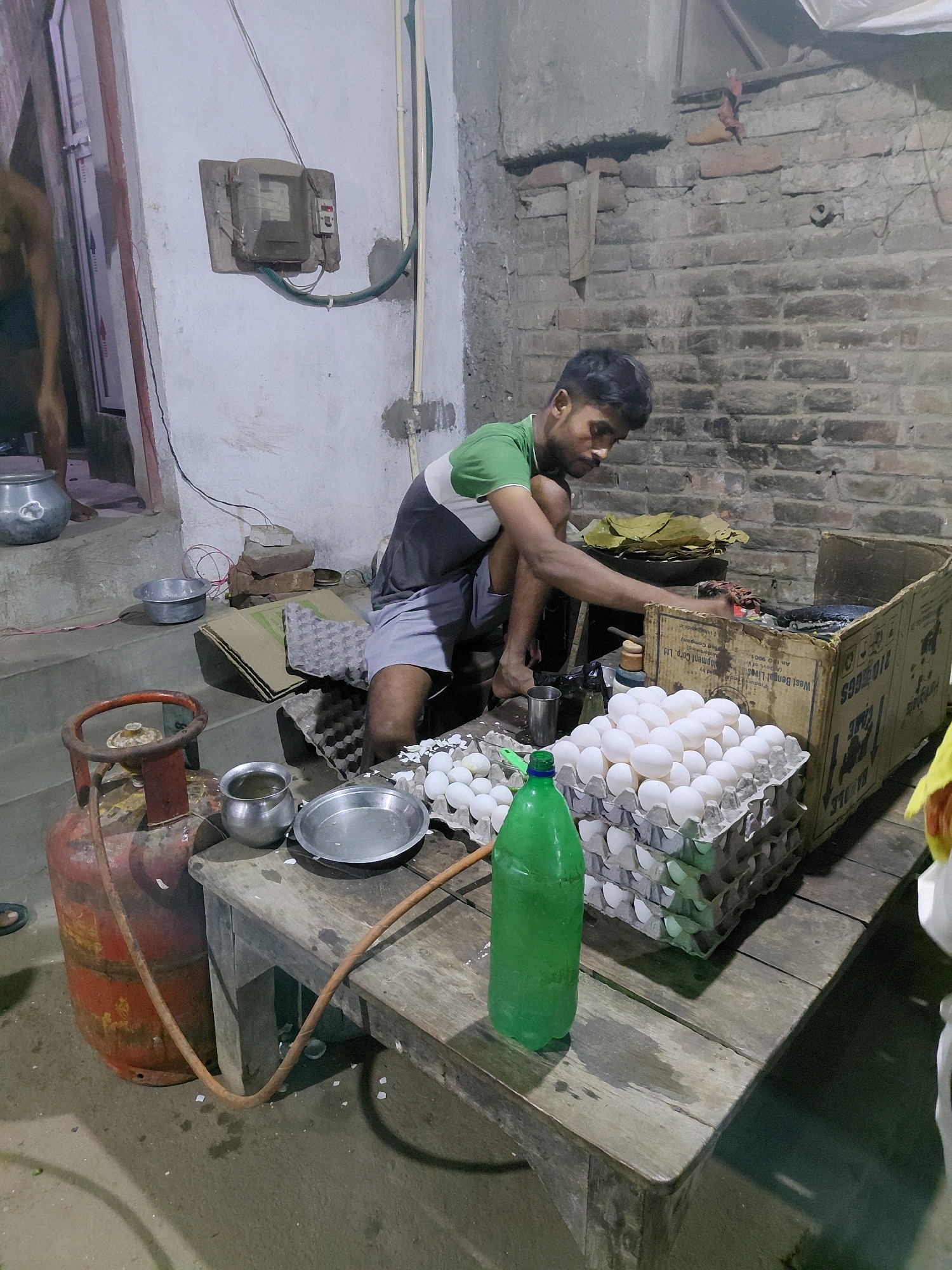 The image depicts an indoor scene, likely a humble dwelling or shop in Patna, India, possibly in a less affluent area given the rustic brick walls and worn wooden surfaces. The focal point is a young man, appearing to be in his late teens or early twenties, dressed in a green and white t-shirt and gray shorts, crouching over a wooden table. He is engaged in the task of handling or preparing eggs, as evidenced by numerous egg cartons filled with white eggs and scattered eggshells on the table.
To the left of the man, a red LPG cylinder with a gas hose connected to a stove (partially visible) suggests cooking activities are common in this space. Several metal bowls and a stainless steel plate are also present on the table, some containing water, further supporting the idea of food preparation. A large green plastic bottle stands upright on the table.
In the background, to the left, a partially visible person is seen standing on a step, next to a large silver pot. An electrical junction box is mounted on a white wall, with wires running across it. The overall environment is dimly lit, with a utilitarian and unrefined aesthetic. The scene conveys a sense of everyday life and labor in a modest setting within Patna.