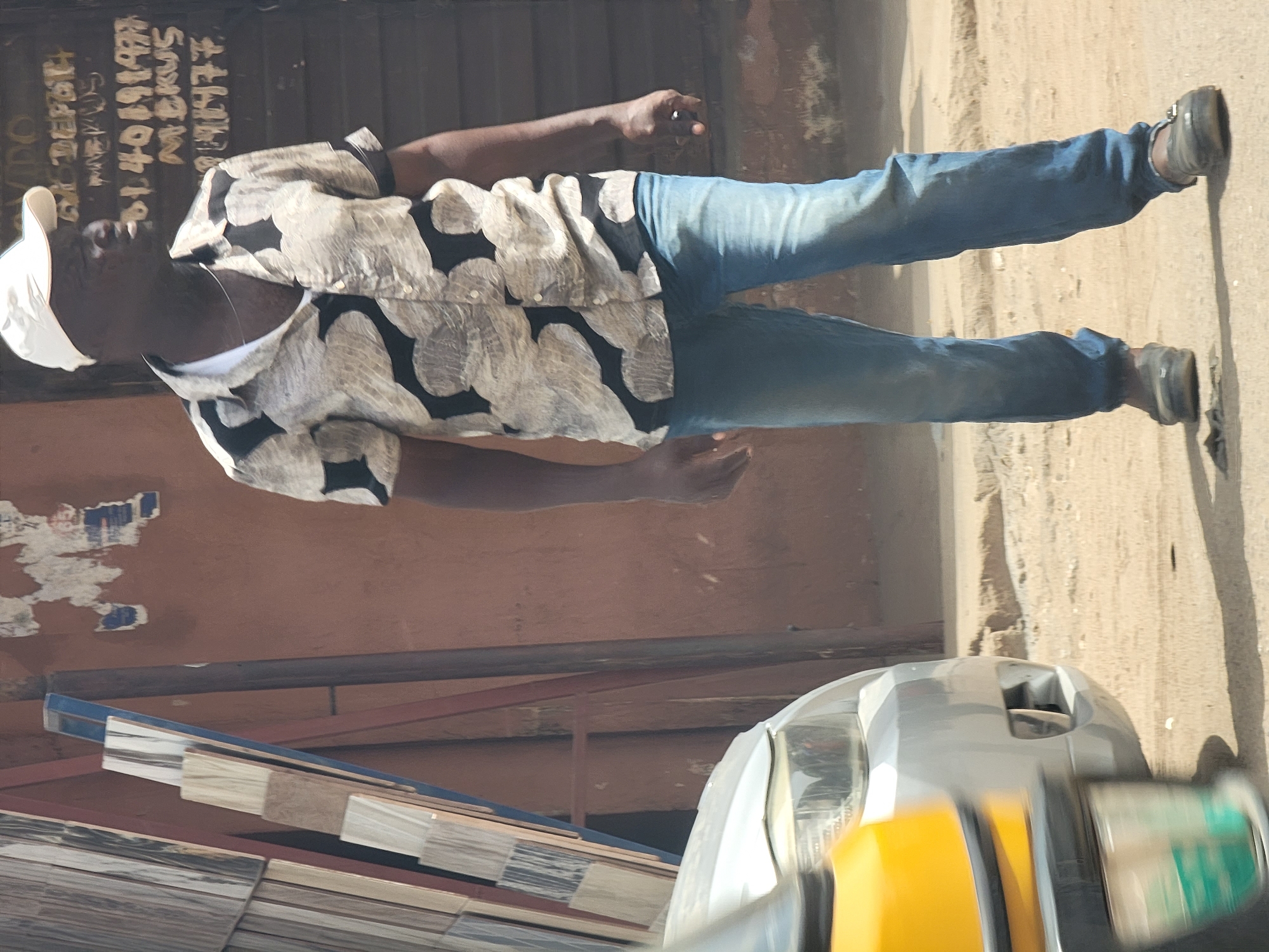 A man wearing a white cap, a patterned shirt, and blue jeans is walking on a sandy surface. He is wearing dark shoes. Part of a silver and yellow vehicle is visible in the lower right corner of the image. The man is in an outdoor setting. The location is Maiburiji, Nigeria, indicated by the writing on the building. Tiled planks are stacked in the bottom left of the scene.