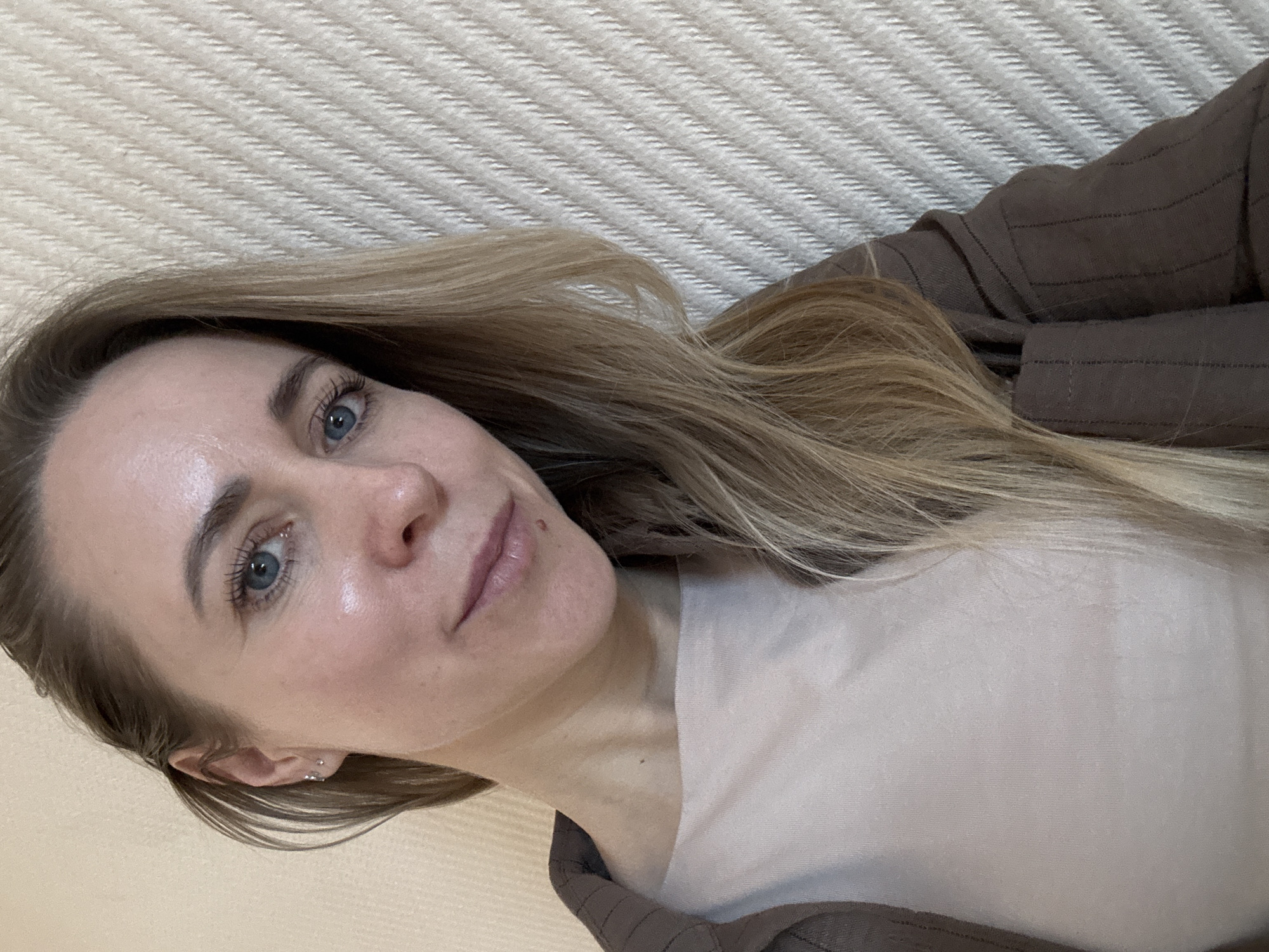 A fair-skinned woman with blonde hair and blue eyes is the primary subject. She is wearing a light-colored t-shirt and a brown striped jacket. Her hair is styled with loose waves. She has a mole on her face near her lip, and the lighting highlights the texture of her skin. The background appears to be a woven textured surface, possibly a wall or a mat. The angle of the shot is slightly canted. There are no discernible landmarks or context clues to indicate the exact location of the image in Chaikovsky, Russia.