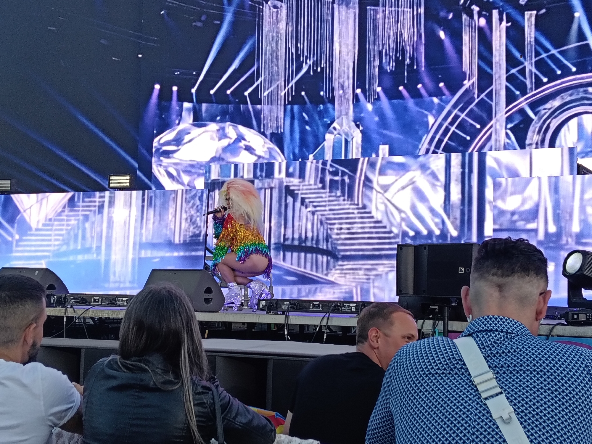 Captured at a venue, possibly in Playa Del Inglés, Spain, an individual with a flamboyant, rainbow-colored outfit and light-colored, teased hair, is positioned on a stage, holding a microphone. The backdrop is an elaborate LED screen display of staircases and diamond shapes with an elaborate lighting display above and behind the stage. In the foreground, several people are seated, viewing the performance. A woman in a black leather jacket has long hair and a man wearing a white t-shirt is seated to her left. Two men are to the right of the frame, one wearing a black shirt and the other in a blue geometric patterned shirt with a white strap across his chest. Two speakers are on the stage near the center of the image.