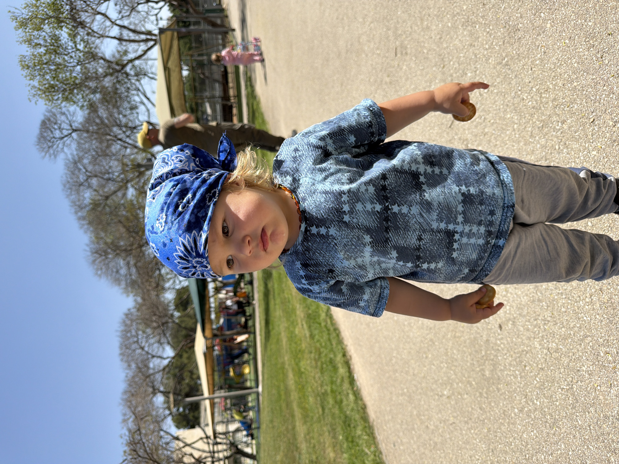 A young child with blonde hair is standing on a paved path outdoors, possibly in a park setting in Kiryat Bialik, Israel. The child is wearing a blue patterned bandana on their head, a blue star-patterned shirt, and gray pants. They are holding a round, flat object in each hand, which may be cookies or crackers. Their right arm is extended, pointing towards something out of frame.
In the background, there is grass, trees, and a partially visible structure that could be part of a playground or park facility. Other individuals are present in the background, including an adult and another child, though they are out of focus. The sky is clear and blue. The photo appears to be taken during the day.