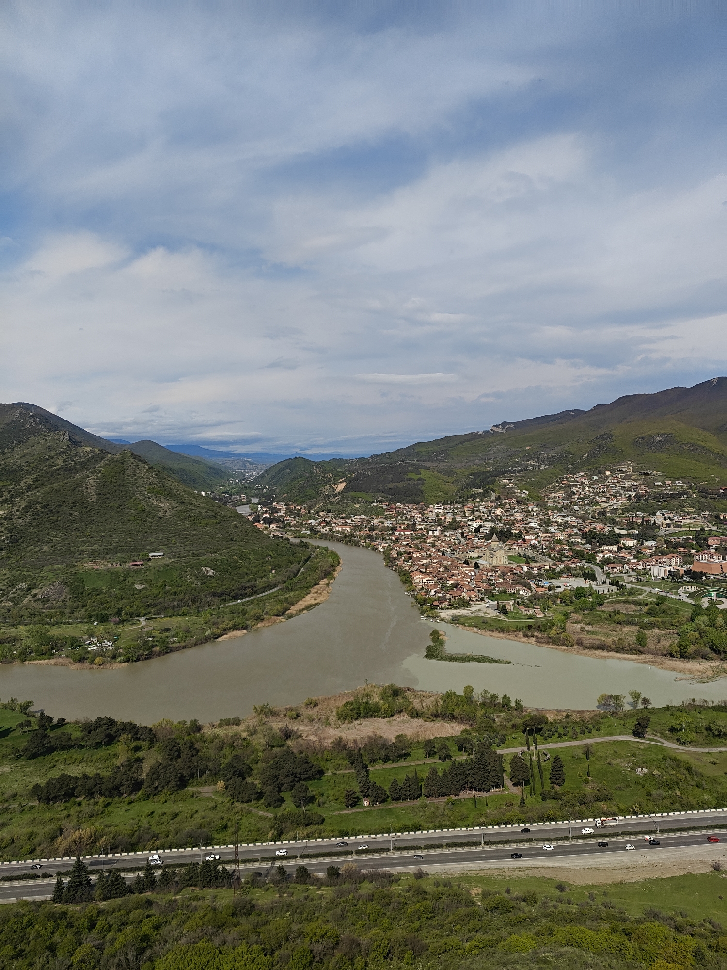 The image shows an aerial view of Mtskheta, Georgia. The confluence of the Aragvi and Mtkvari rivers is prominent, with the town nestled between the rivers and surrounding hills. The Mtskheta skyline features traditional red-tiled roofs of buildings. A multi-lane highway runs along the lower edge of the image, with cars visible. The landscape is green with trees and vegetation. The sky is partly cloudy.