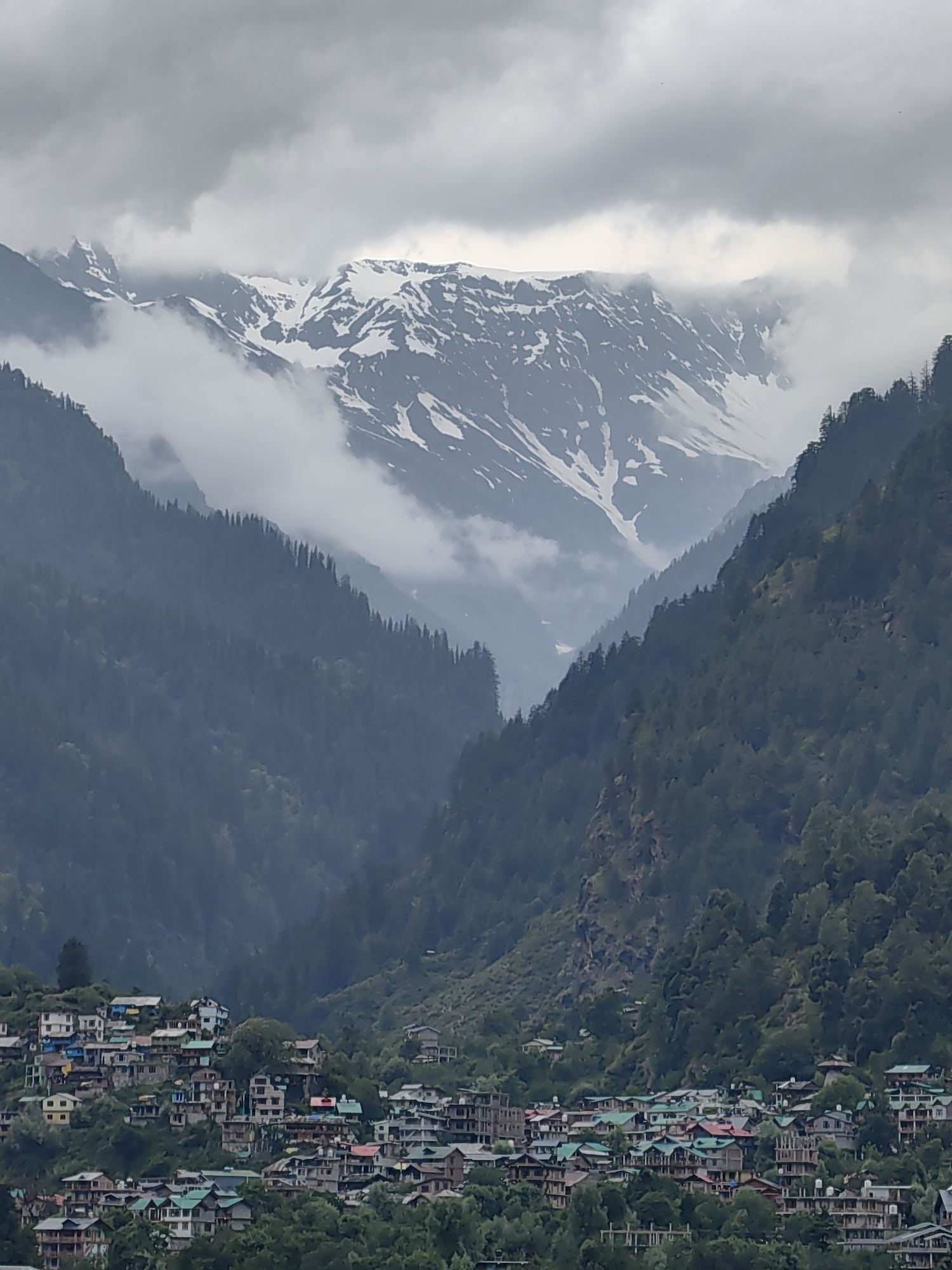 This is a vertical, eye-level shot of the town of Vashisht, India, nestled in a valley surrounded by lush green mountains. In the background, snow-capped peaks rise majestically, partially obscured by soft, wispy clouds. The town itself is a dense collection of multi-story buildings with brightly colored roofs, scattered across the steep hillside. Trees and vegetation are interspersed among the buildings and cover the surrounding slopes, giving the impression of a vibrant community integrated with nature. The sky is overcast, with dramatic clouds adding to the mountainous landscape's mood. There are no visible people or animals in the frame.