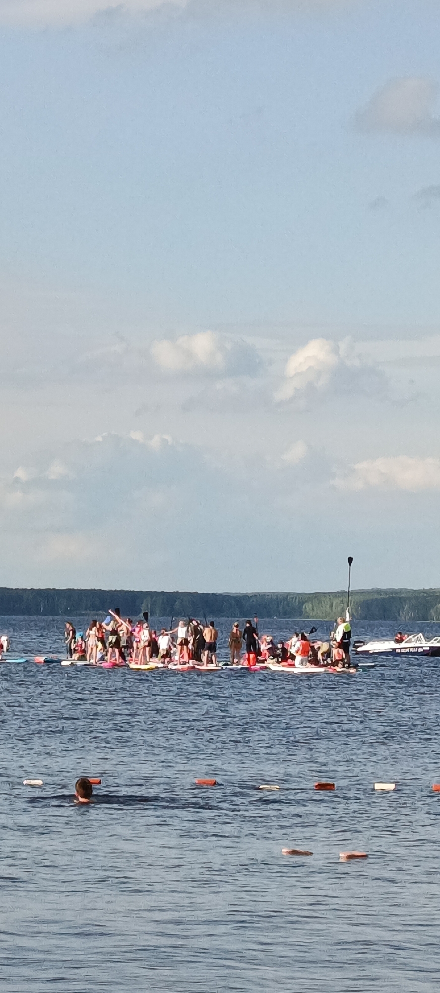 The image depicts a gathering of people on stand-up paddleboards in a body of water, likely the Kama River near the city of Chaikovsky, Russia. The sky is partly cloudy with a light blue hue. In the foreground, a person is swimming with their head and shoulders above the water. Further out, a large group of people are standing on numerous paddleboards, many in swimwear, some holding paddles. The group appears to be engaged in a recreational activity or event. In the background, a line of trees forms the shoreline, and a small boat is visible to the right. The water is calm with visible ripples.