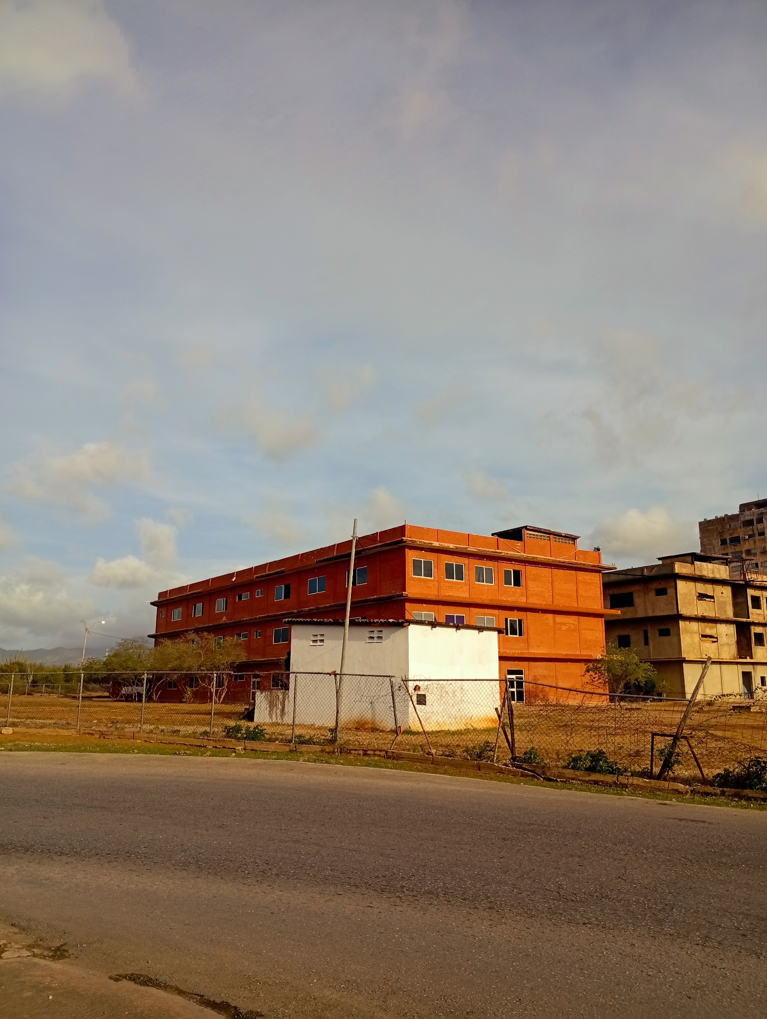 This is a medium-shot, eye-level, outdoor, vertical photograph of an abandoned construction site in Juan Griego, Venezuela. In the foreground, a paved road is visible, with a curb and a strip of dry grass. A chain-link fence runs along the side of the road, partially obscuring the view of the buildings behind it.
The main subjects are two unfinished buildings. The building on the left is a large, rectangular structure with an orange brick facade. It has multiple floors with numerous rectangular windows, some of which are boarded up or appear to be missing glass. There is a small, white, single-story building in front of the orange building, also with windows.
To the right of the orange building, another multi-story building is visible. This building has a concrete exterior and appears to be in an even more advanced state of disrepair, with visible structural damage, missing walls, and broken windows. A taller, darker building, possibly a skyscraper, is visible in the background to the far right, also appearing to be under construction or abandoned.
The environment is arid, with dry, yellowish grass and sparse, scrubby trees. In the distance, mountains are visible under a cloudy sky. The lighting suggests it is daytime, likely late afternoon, with soft shadows and warm tones. There are no people or animals visible in the image. The overall impression is one of decay and abandonment.