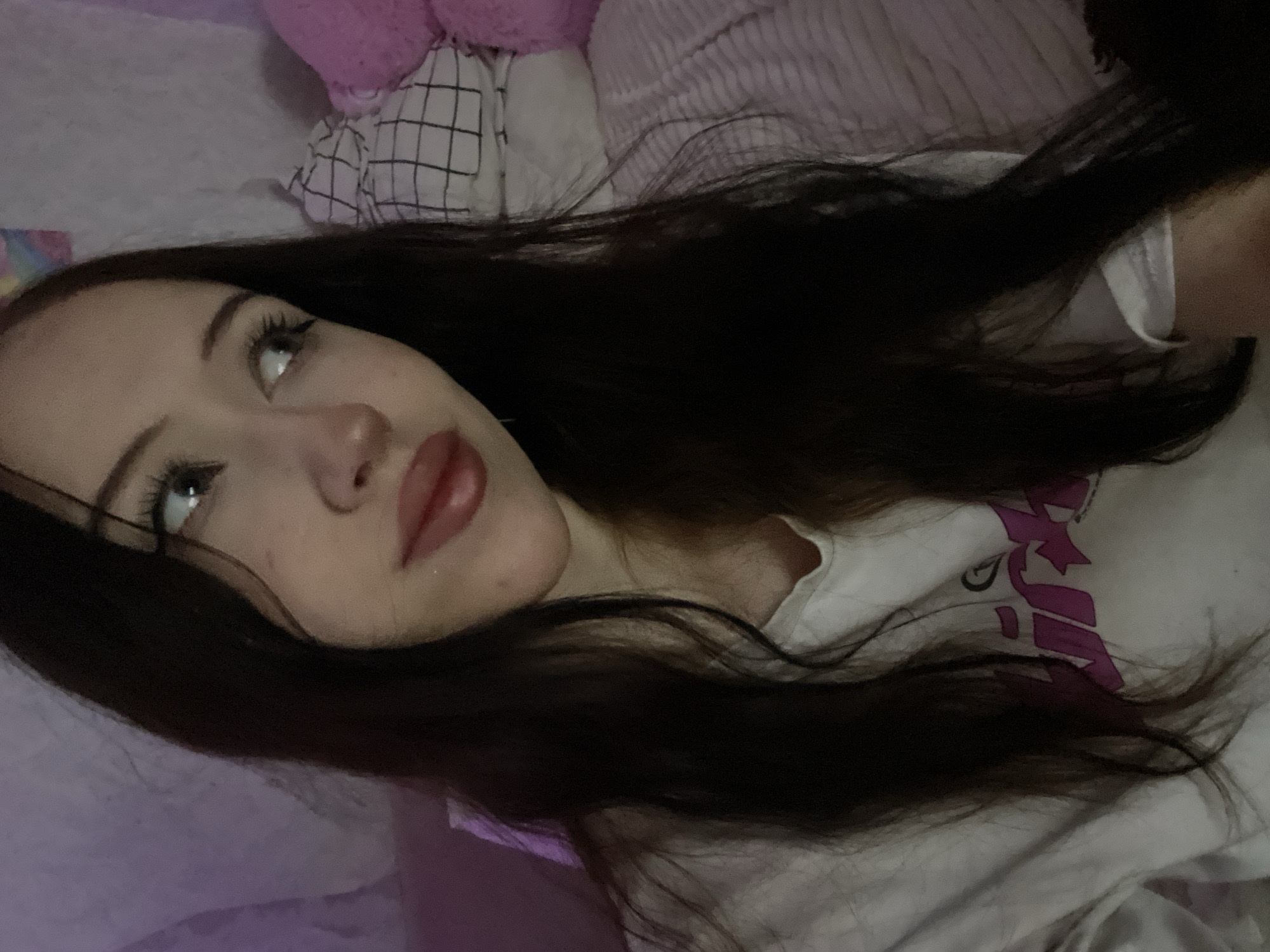 The image shows a close-up of a young woman with long, dark hair. She is wearing a white t-shirt with a pink graphic on it. Her eyes are looking to the right, and her lips are slightly parted. She appears to be indoors, possibly in a bedroom, with soft lighting and some plush toys in the background. There is no indication of her location within Votkinsk, Russia.