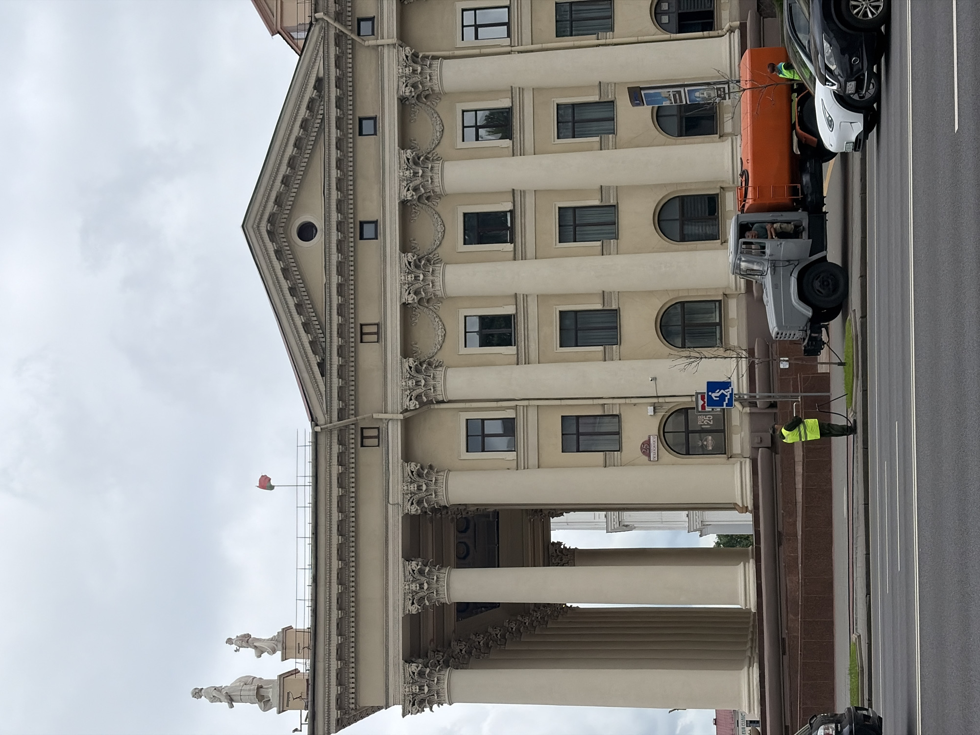 The image captures a street view in front of a grand, neoclassical building in Minsk, Belarus. The building features a prominent colonnade with large, ornate columns. Above the colonnade, there are statues and a flag. The sky is overcast.
In the foreground, a grey truck with an orange tank is parked on the side of the road. A person in a high-visibility vest is standing next to the truck. Several cars are also visible on the road, including a white electric vehicle and a black SUV. The street is asphalt, and there is a sidewalk and a grassy area between the road and the building. A signpost with a blue pedestrian symbol and an arrow is also present.
The building's facade is decorated with classical architectural elements, including entablatures, friezes, and arched windows. The street name sign visible on the building reads "25 Независимости" (25 Nezalezhnosti), indicating the address on Nezalezhnosti Avenue in Minsk. This avenue is a major thoroughfare in the city.