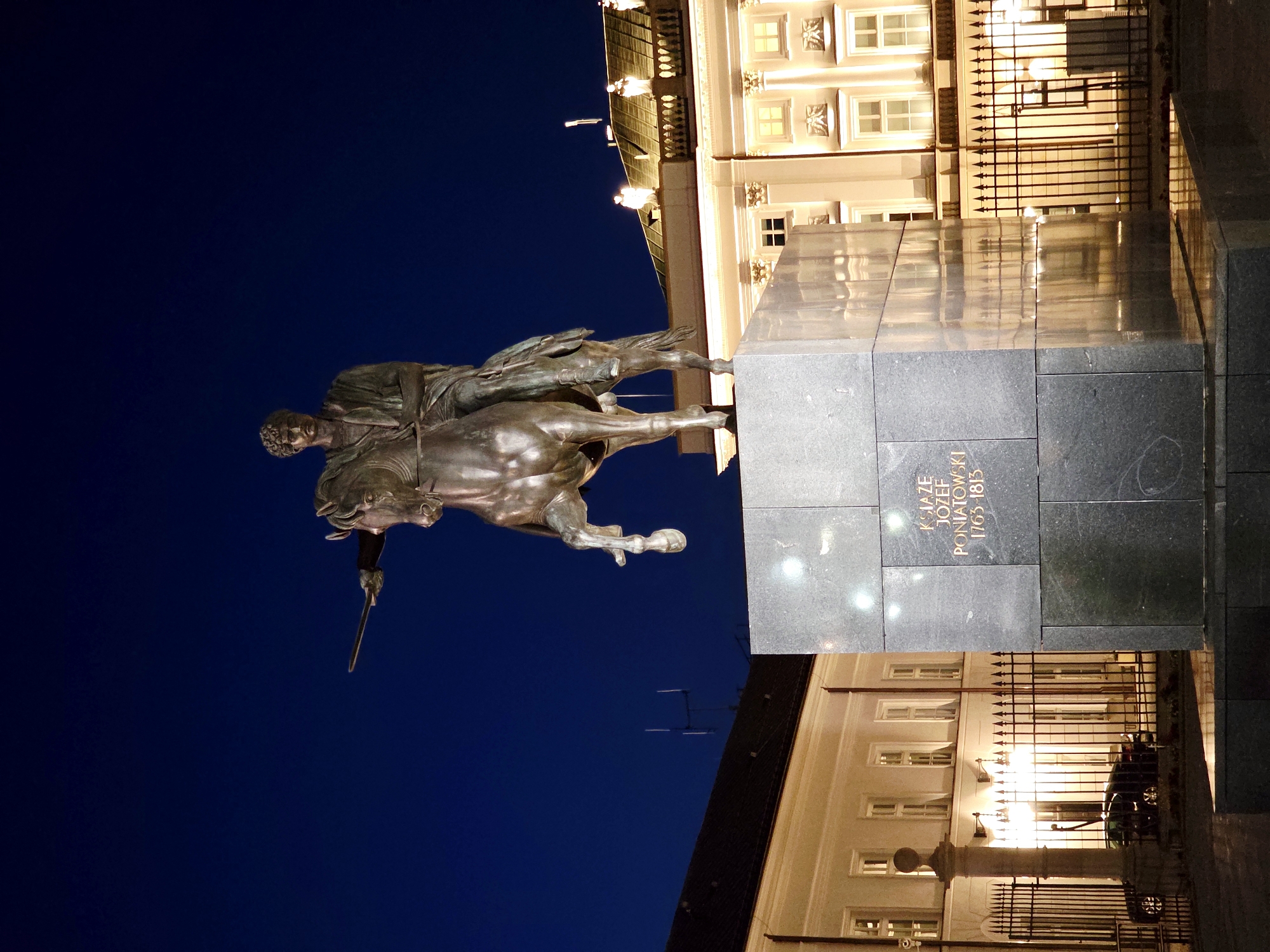 This is a nighttime, low-angle, full-shot photograph of the Jozef Poniatowski monument in Warsaw, Poland. The monument, made of bronze, depicts a man on horseback, pointing forward with a sword. The man wears a toga-like garment. The horse is in mid-gallop, with its front legs in the air. The monument stands on a large, dark granite pedestal with gold lettering on one side that reads "KSIAZE JOZEF PONIATOWSKI 1763-1813." In the background, a white neoclassical building with illuminated windows and decorative elements is visible. A black metal fence runs in front of the building. A dark-colored car is parked behind the fence. The sky is a deep, clear blue. The image is well-lit, with the monument and building brightly illuminated by artificial lights.