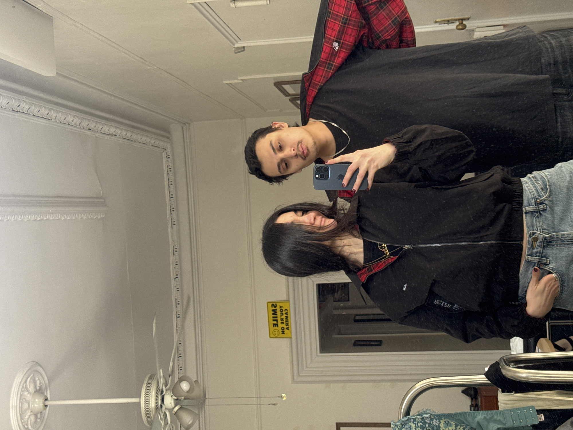 A mirror selfie shows two individuals in a room. The person on the left wears a black t-shirt with a red plaid flannel shirt underneath and a silver chain. The person on the right wears a black jacket with red plaid trim. They are holding a blue iPhone. The room has a ceiling fan and what appears to be window frame and moulding characteristic of older buildings in New York City. The environment includes objects, a mirror, and window frame. This is an indoor scene with elements reminiscent of an older apartment building interior typical of New York City. The action is taking a photograph, the interaction being a couple.