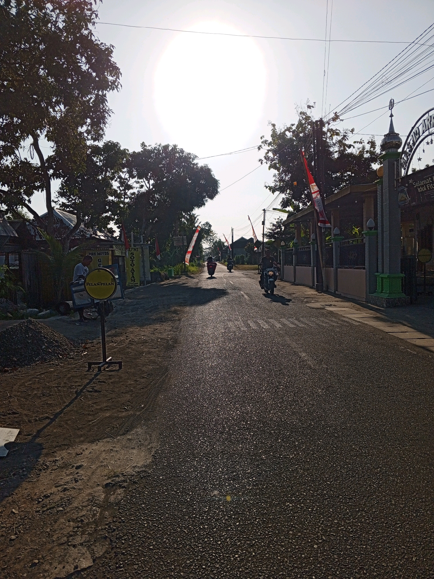 The image depicts a street scene in Bantul, Indonesia, bathed in the bright sunlight of what appears to be late afternoon or early morning, judging by the low sun angle casting long shadows. The road is asphalt, with visible tire tracks on the shoulder in places. On the left side of the road, there is a pile of gravel and a yellow circular sign on a stand with the Indonesian word "PELAN-PELAN" (slowly) visible. A man in a white t-shirt stands near the sign, looking at it or something behind it. Further down the road, several motorcycles are in motion. On the right side of the road, there is an entrance gate with Islamic architectural elements, including a dome and minaret-like structures. Banners with Indonesian text and red and white flags are displayed along the street and near the gate. Buildings with visible roofs and walls line the sides of the road. Trees with dense foliage are also present. Power lines crisscross the sky.