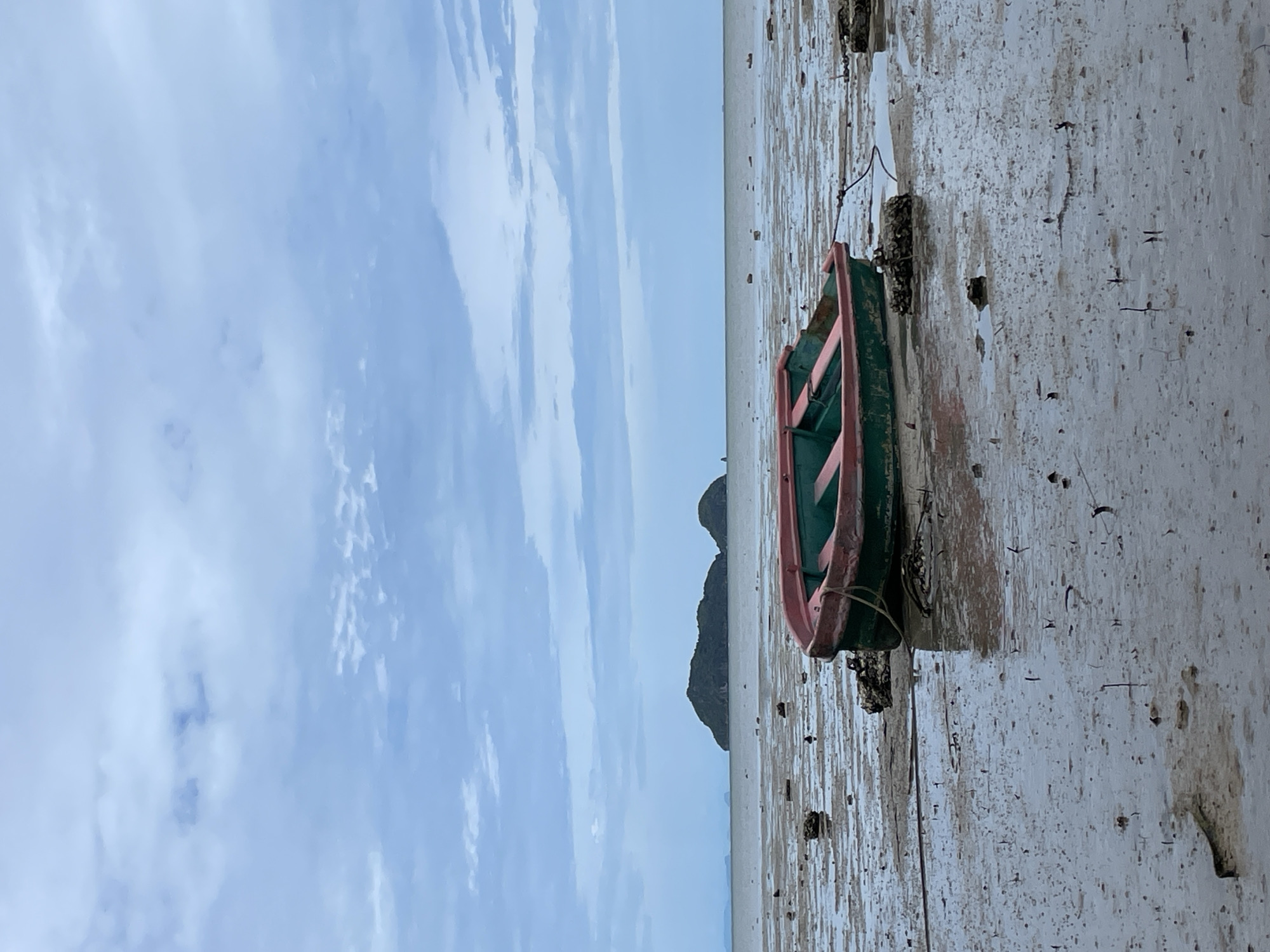 A small, green and pink boat sits beached on a muddy shoreline in Baan Sri Thanu, Thailand. The boat's hull rests on the wet sand, with a rope attached. Beyond the beach, a small, dark, rocky island is visible in the distance. Above the island, a light blue sky is streaked with thin, white clouds. The foreground mud is textured with grooves and small debris.