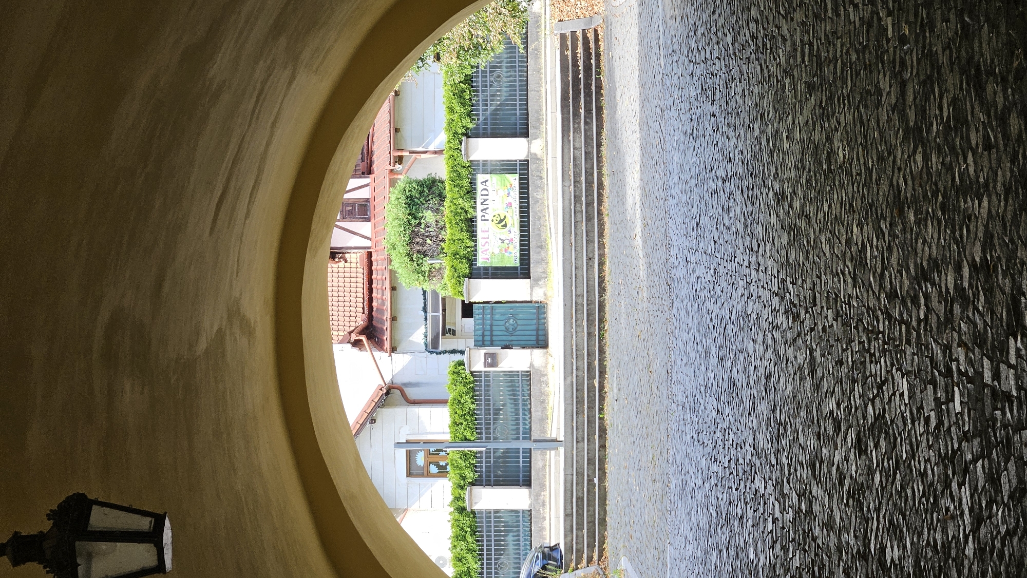 The image captures a view through a stone archway, looking out onto a street in Prešov, Slovakia. In the foreground, the cobblestone street is wet, suggesting recent rain, and reflects the light. Beyond the street, a set of stone steps leads up to a paved walkway. A black metal fence runs along the walkway, behind which is a low hedge. A green metal gate is visible within the fence. To the right of the gate, a large banner with "JASLE PANDA" and cartoon pandas is displayed. Behind the fence and hedge, a white building with a red-tiled roof and a half-timbered upper section is visible. A dark, traditional-style lantern hangs from the archway on the left side of the image. The overall scene is brightly lit by sunlight.