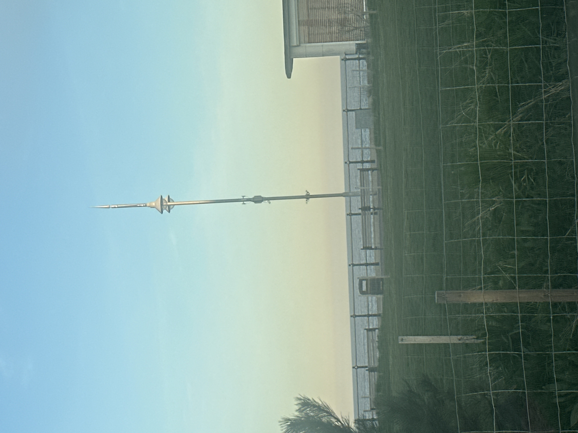 The image depicts a view in East Suffolk, United Kingdom. The seaside is the main focus, observed through a wire fence. A tall metal structure stands in the distance. Near the fence, wooden planks are visible amidst greenery. The sky transitions from blue to yellow-white near the horizon. The scene includes a building and the sea, with beach furniture visible along the coastline.