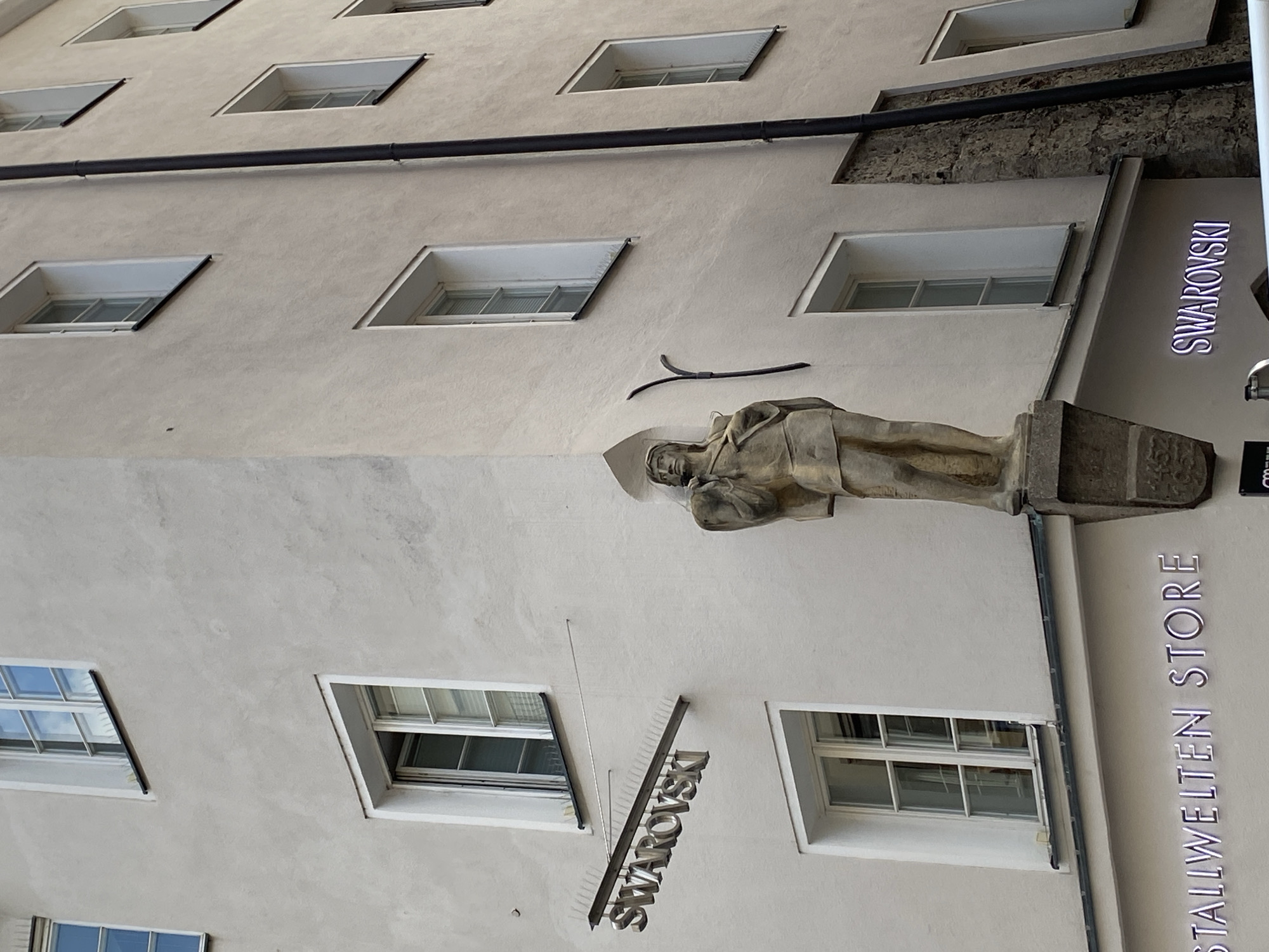 The image shows the corner of a building in Innsbruck, Austria, with a statue of a man in the foreground. The statue is made of stone and depicts the man wearing a tunic and holding a staff. The building is light-colored and has several windows. A sign for "SWAROVSKI" is visible on the building. Below that, another sign reads "STALLWELTEN STORE". The statue is positioned on a ledge protruding from the building. The overall scene appears to be an urban streetscape.