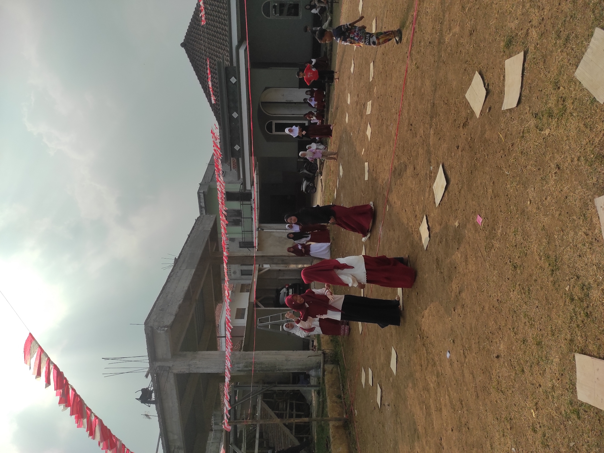 In Sumedang, Indonesia, a group of children and adults are gathered outdoors in a dirt and grass area. The scene appears to be part of a community event or celebration, possibly related to Indonesian Independence Day, given the red and white decorative banners strung across the area and draped over buildings. In the foreground, several pieces of cardboard are scattered on the ground, suggesting they may be part of a game or activity.
Several individuals, predominantly young people, are engaged in some form of organized activity. A few are wearing traditional Muslim attire, including headscarves (hijabs) in various colors, and long skirts or pants. Some of the women are dressed in maroon and white outfits. A young boy in a colorful shirt and patterned pants is walking in the foreground.
In the background, there are unfinished and completed structures. One building, under construction, features exposed concrete beams and rebar. Another building has a traditional tiled roof and visible windows. The sky is overcast with scattered clouds. The overall atmosphere suggests a lively outdoor gathering for children and the community.