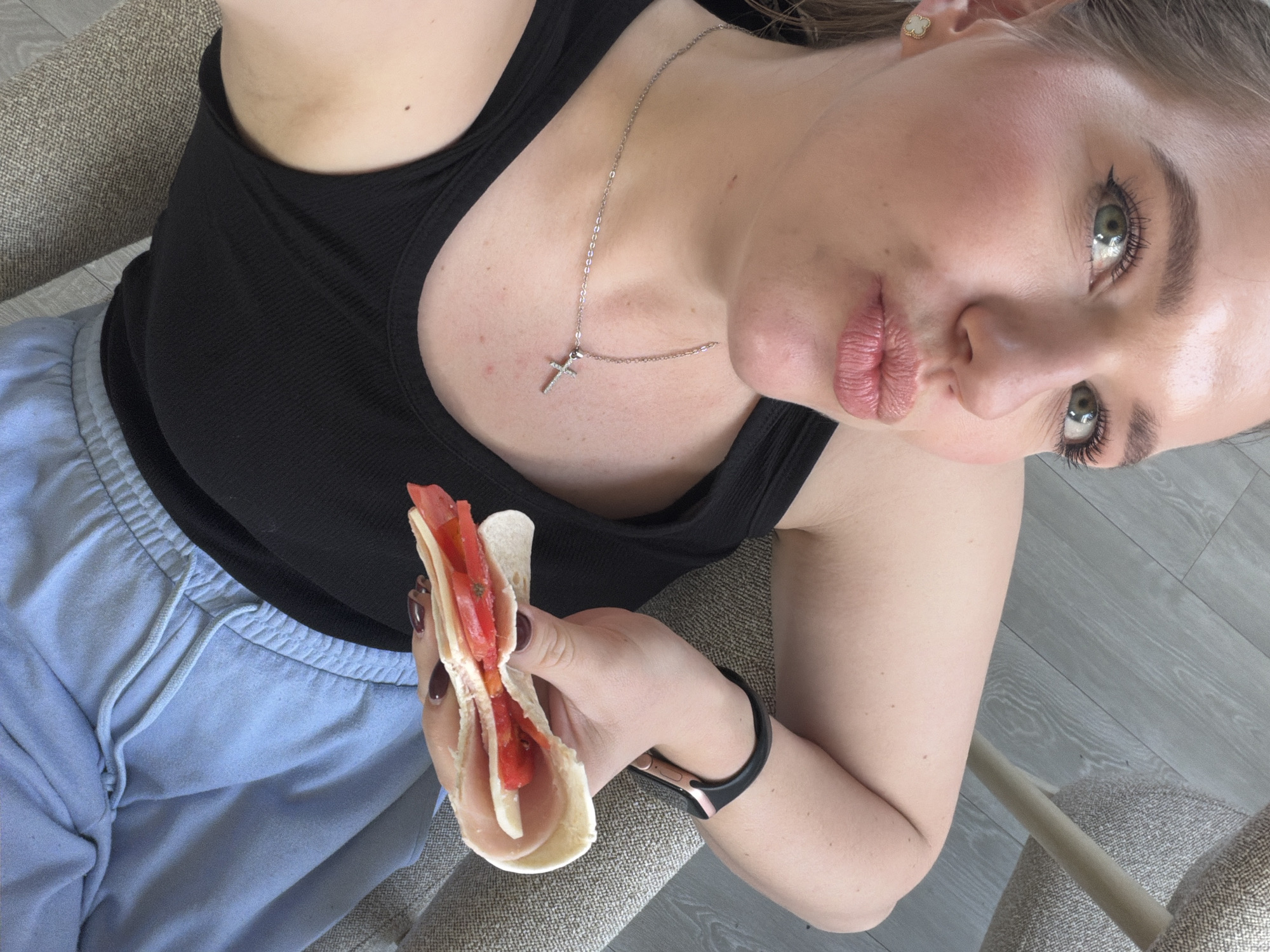 A woman is taking a selfie while holding a sandwich. The sandwich contains tomato and cheese. She is wearing a black tank top and light blue sweatpants. She has a silver cross necklace on. She is seated in an upholstered chair. The floor appears to be grey wood-look laminate. It cannot be determined if the location is Dreiliņi, Latvia, from the image.