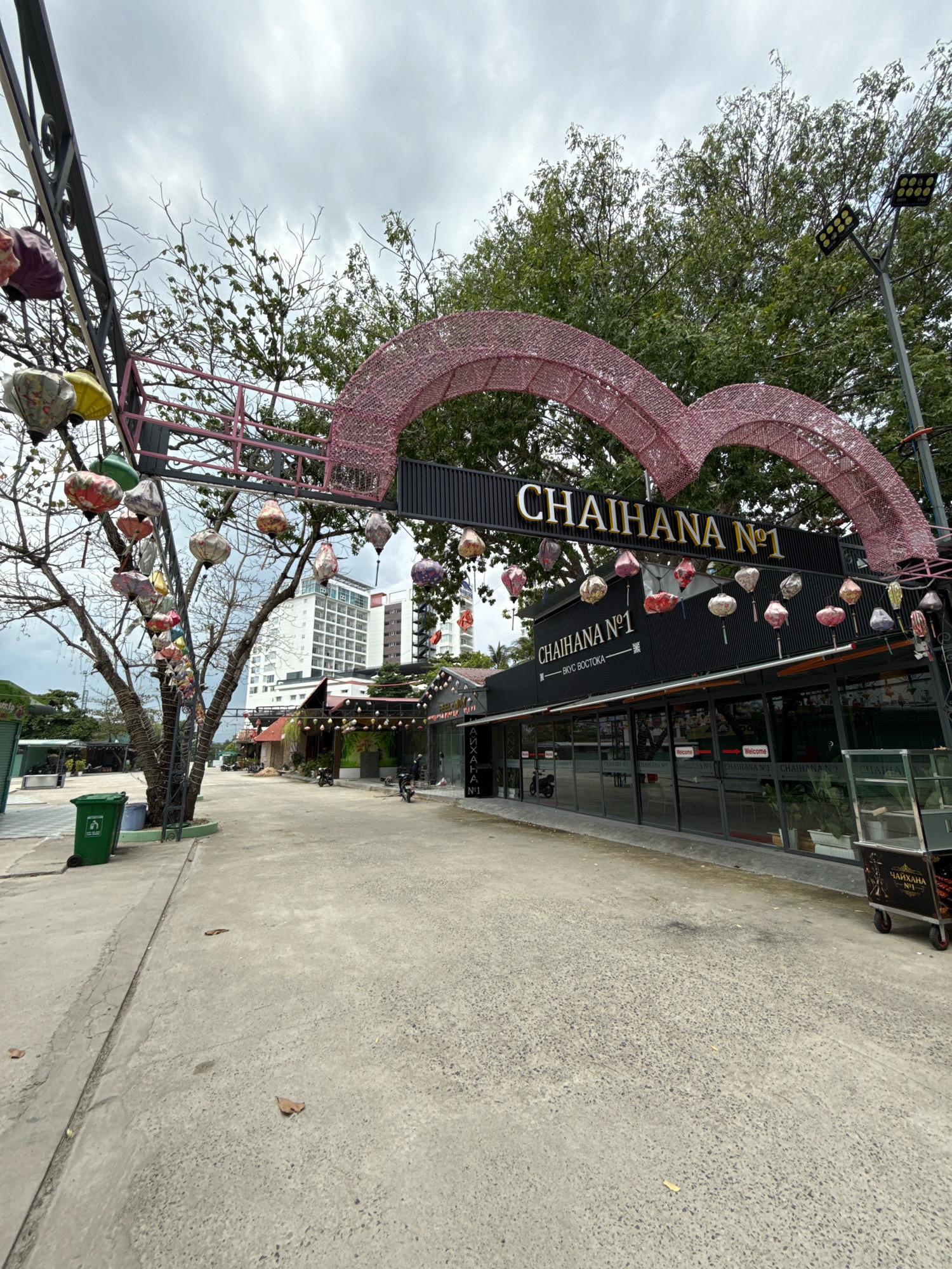 The image captures an outdoor scene in the city of Nha Trang, Vietnam, likely an entertainment or dining district. In the foreground, a concrete pathway leads towards a building adorned with a large, pink, heart-shaped archway. The building itself is modern, with a dark facade and large glass windows, and bears the signage "CHAIHANA N°1" in prominent lettering. This establishment appears to be a cafe or restaurant.
Suspended from a dark metal structure and a nearby tree are numerous colorful, decorative lanterns, adding a festive ambiance to the scene. A tall, slender building, characteristic of urban Vietnamese architecture, stands in the background, partially obscured by trees and the sky. The sky is overcast, suggesting a cloudy day.
To the left of the pathway, a bright green trash bin is visible. In the middle ground, several motorcycles are parked, indicating a place frequented by locals or tourists. The overall environment suggests a commercial area with a mix of modern and decorative elements. The weather appears calm, with no significant wind or precipitation. The image composition focuses on the entrance of "CHAIHANA N°1," drawing attention to its unique architectural features and decorative elements.